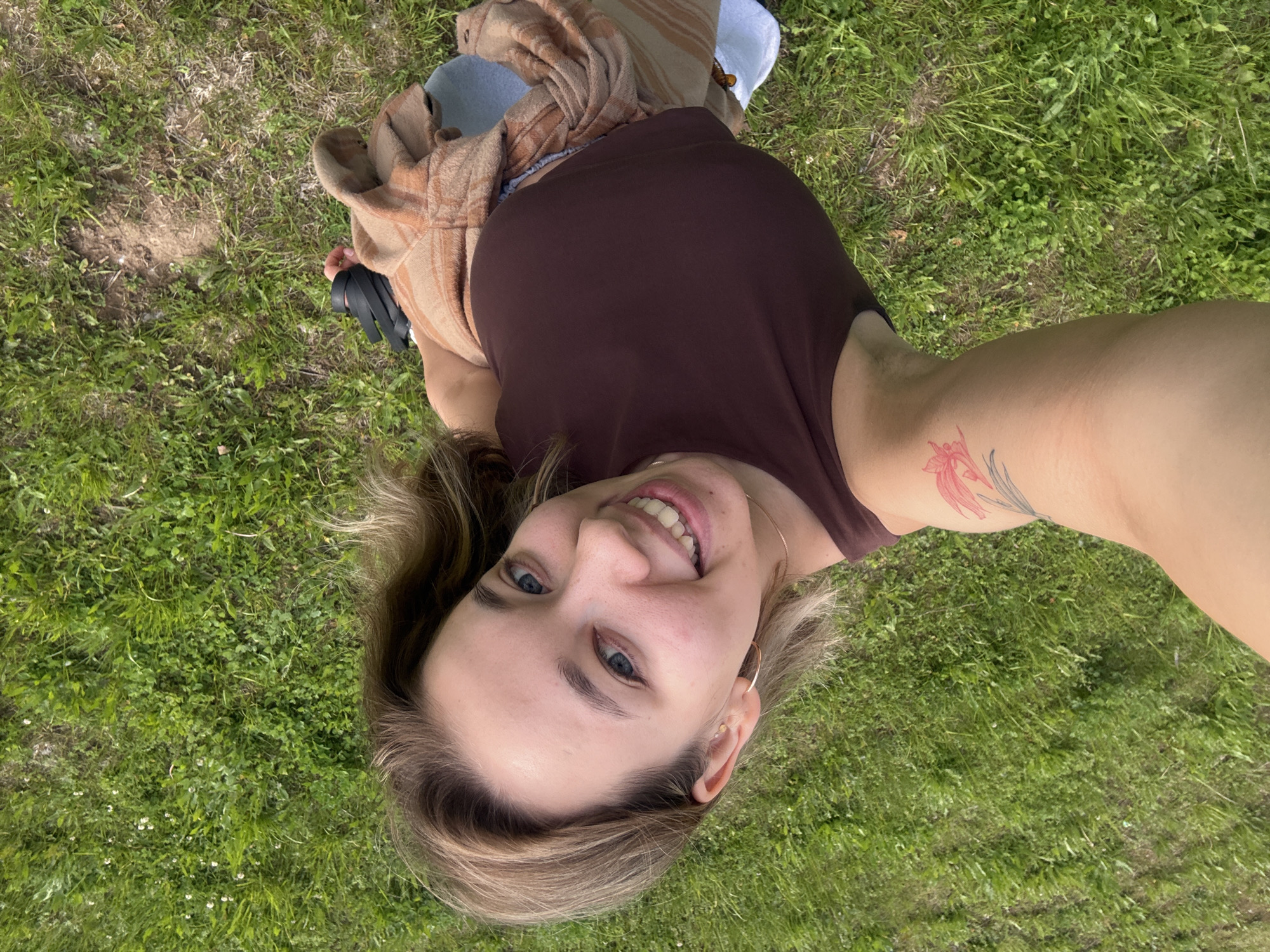 A young woman is captured in a top-down perspective, lying on her back on a lush green grassy surface. She looks directly at the camera with a broad, joyful smile, revealing her teeth. Her blue eyes are bright, and her light-colored hair, a mix of blonde and light brown, frames her face.
She is dressed in a dark brown tank top. A light brown and orange striped garment, possibly a shirt or blanket, covers her lower torso and legs, which are mostly out of frame. A light blue fabric item is visible underneath the striped garment, suggesting she might be resting on a blanket or cushion. A pair of dark-colored sandals are partially visible near her left side.
On her right upper arm, a prominent tattoo features a stylized floral or plant design in red, accented with black or grey details. She accessorizes with a thin gold chain necklace and small hoop earrings.
The setting is an outdoor grassy area, predominantly covered in vibrant green blades of grass, with some small white flowers and patches of drier ground visible. The lighting appears bright and natural, indicating it is daytime. The image captures a moment of contentment and relaxation, likely in an outdoor space in Chaikovsky, Russia. There is no visible text in the image.