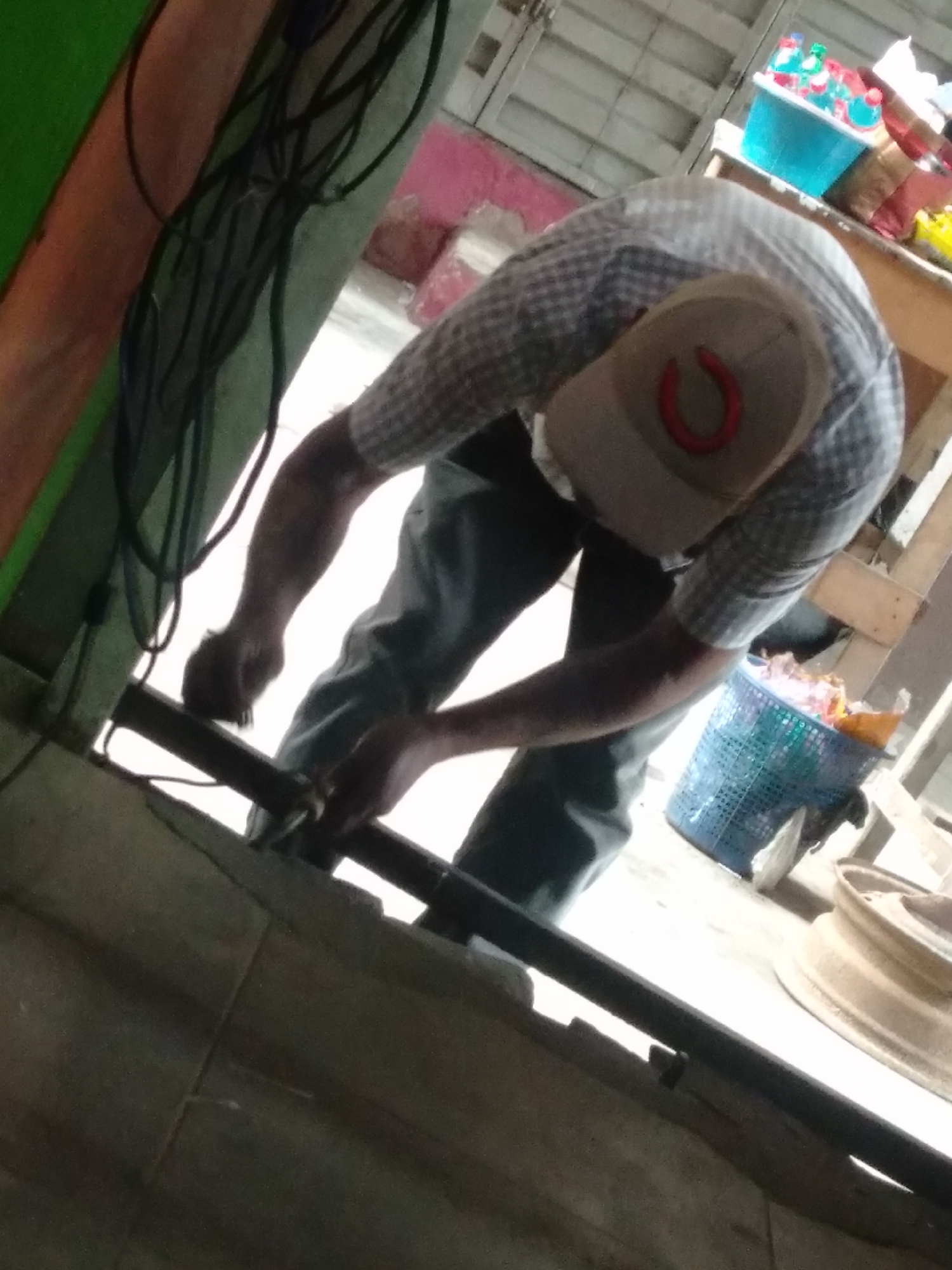 In the city of Shangisha, Nigeria, a person is shown bent over a dark-colored pipe or rod that runs horizontally across the foreground. The individual wears a checkered shirt and blue jeans, and a light-colored baseball cap with a red horseshoe logo. Their hands are occupied with the pipe, suggesting an action related to maintenance or construction. Behind the person, a blue plastic basket filled with various items, including what appears to be trash bags, is visible. In the background, a metal ceiling structure and shelves with containers can be observed. The overall scene suggests an outdoor or workshop environment.