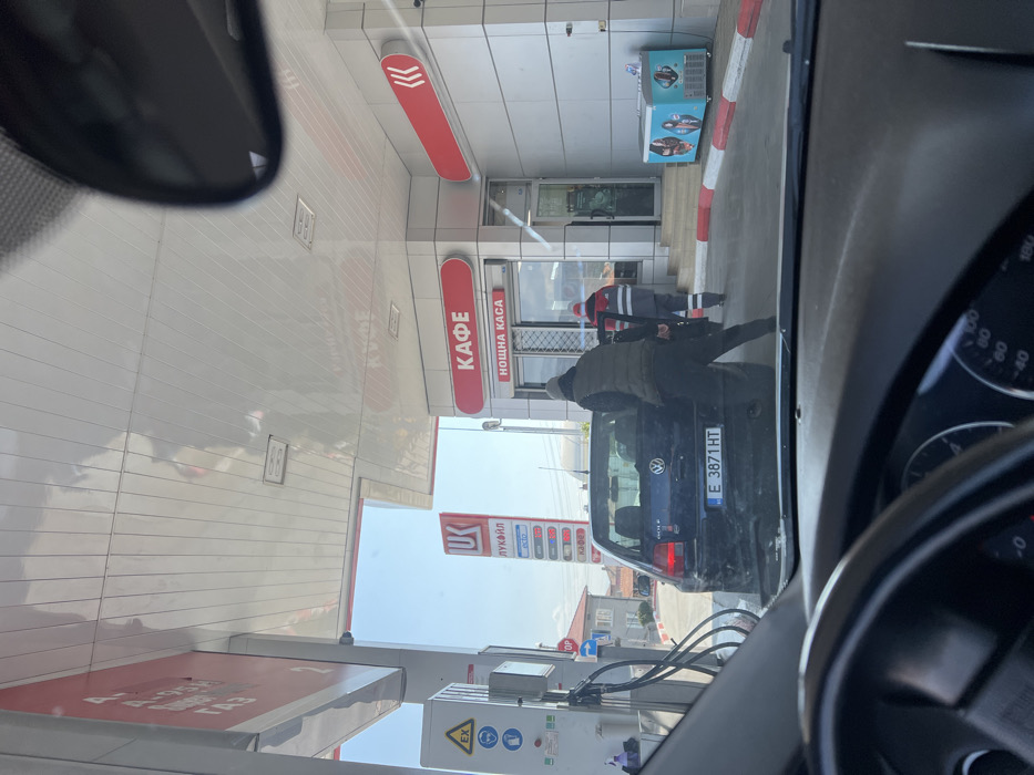The image depicts a daytime scene at a Lukoil gas station in Banichan, Bulgaria, viewed from the interior of a vehicle. A dark blue Volkswagen Golf hatchback, bearing Bulgarian license plate E 3871 HT, is positioned at a fuel pump. A male individual, wearing a dark beanie, olive green puffer vest, and dark pants, is bent over at the rear of the Volkswagen Golf, appearing to interact with its trunk. A second male, dressed in a red and white work uniform, walks towards the entrance of the gas station building.
The gas station facility features a white and grey fuel pump structure with hoses extended towards the Golf. A tall Lukoil totem sign, displaying fuel prices in Bulgarian Cyrillic, is situated behind the Golf. The gas station building is a light grey paneled structure with red accents, marked with signs reading "КАФЕ" (Cafe) and "НОЩНА КАСА" (Night Cashier) above a barred window. An aqua blue freezer unit is located on a raised platform with red and white striped curbing near the building's entrance. A large, white slatted canopy covers the fueling area. In the background, residential buildings with red-tiled roofs and sparse vegetation are visible under a clear blue sky. The immediate foreground reveals portions of a steering wheel and dashboard from the observer's vehicle.