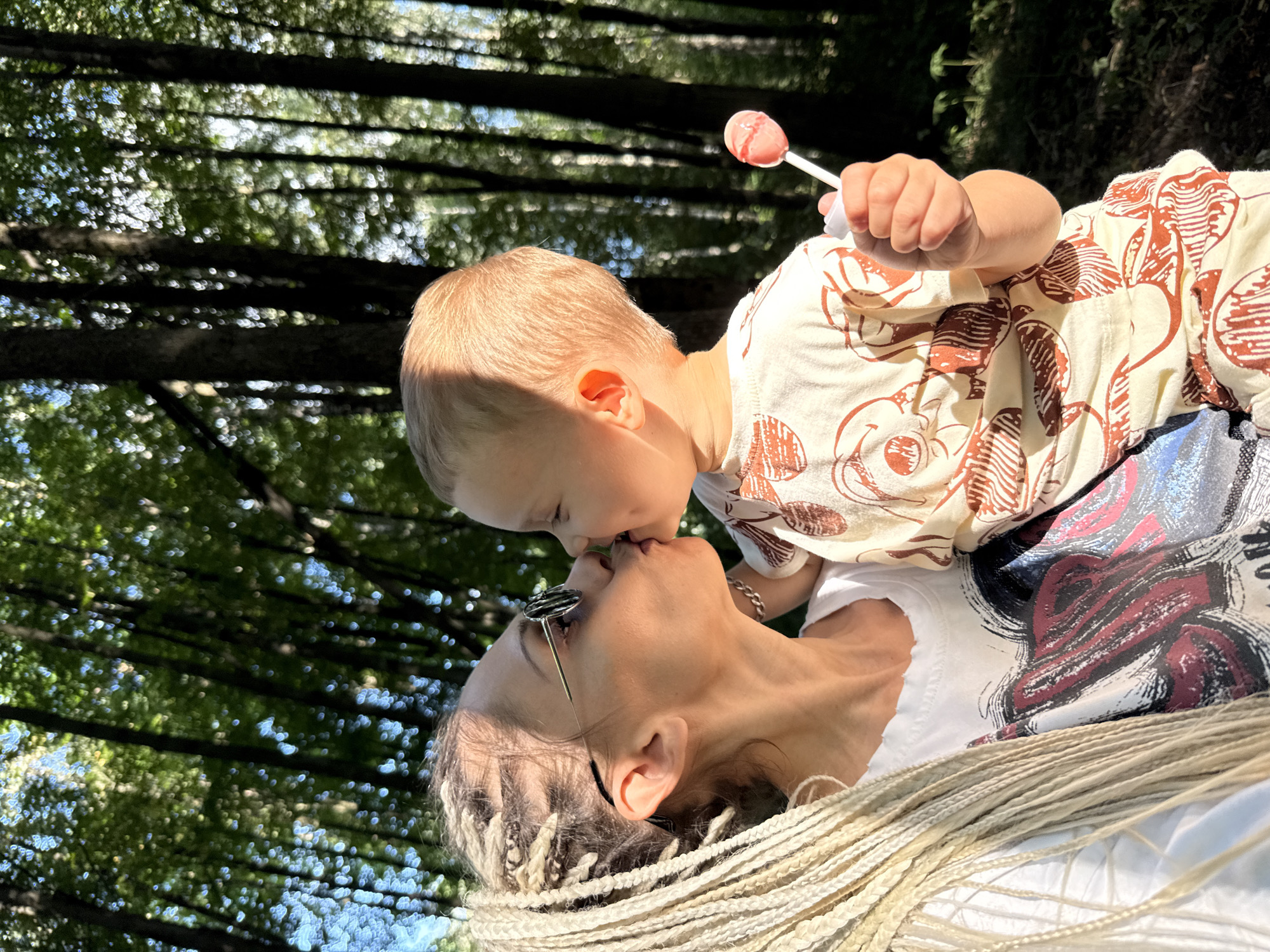In this close-up, low-angle outdoor shot taken in Rusanovo, Russia, a mother and her young child share a tender moment. The mother, with blonde braids and round sunglasses perched on her nose, looks up to kiss her baby, who is being held aloft. The baby, with fair hair, is reaching out with a pink lollipop in one hand. Both are dressed casually, the baby in a light-colored outfit with a repeating pattern of what appears to be Mickey Mouse faces, and the mother in a white t-shirt. The background is dominated by the dappled sunlight filtering through the dense canopy of trees, suggesting a midday or early afternoon setting on a clear day. The out-of-focus green foliage and sunlight create a soft, natural ambiance. The image evokes feelings of warmth, love, and the simple joys of childhood.