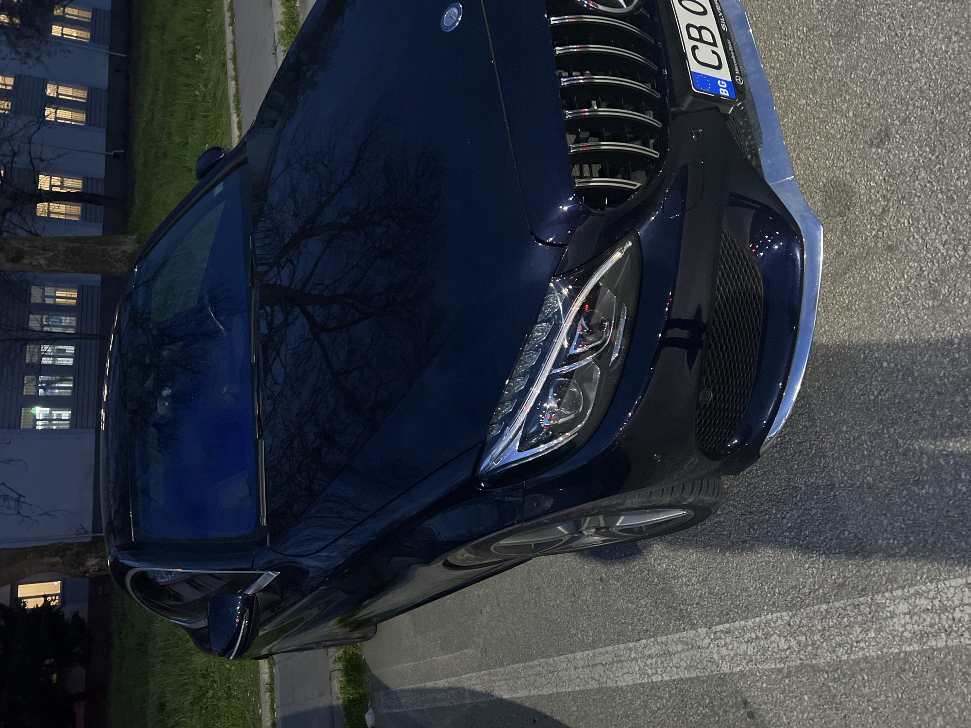The image depicts the front end of a dark-colored Mercedes-Benz car parked on a street. The car has a Bulgarian license plate reading "CB 0". The front grille is prominently visible, along with the driver's side headlight. The car is parked beside a curb and next to a grassy area. In the background, there is a multi-story building with illuminated windows. Based on the license plate and general aesthetic of the area, it is likely the location is in Bulgaria, but the specific location in Plovdiv cannot be determined from this image alone. No people are visible in the frame. The image appears to have been taken at night or in low light conditions.