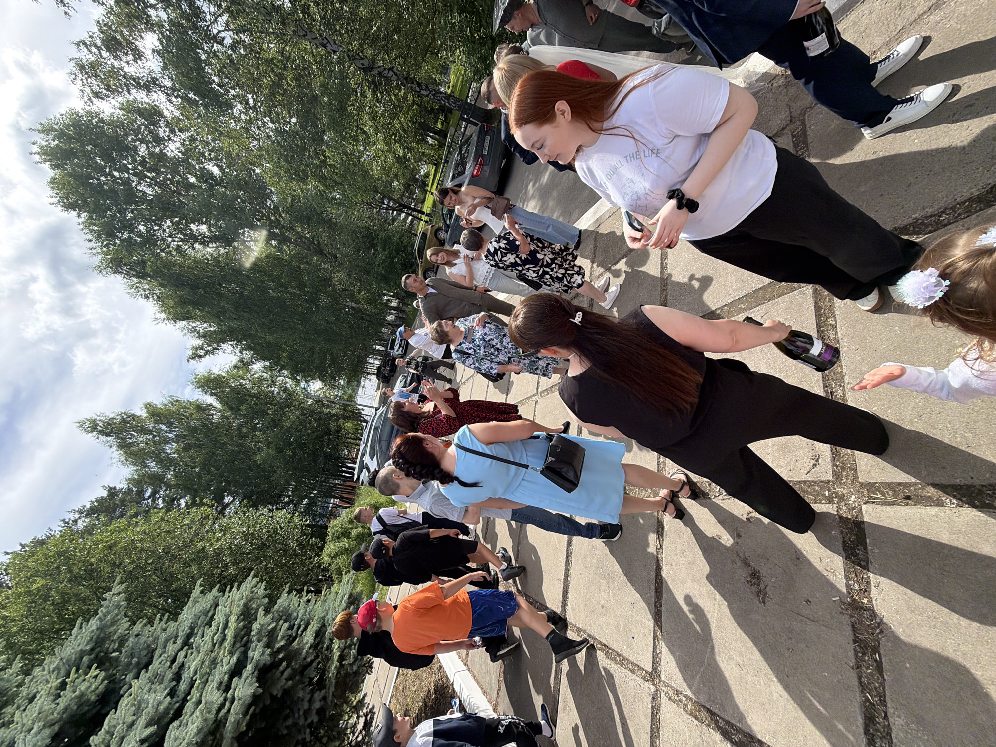 In Chaikovsky, Russia, on a sunny day, a group of people are gathered outdoors on a paved area lined with trees. The setting appears to be a park or a residential area, with the overcast sky suggesting late afternoon. Several individuals are in conversation, while others are walking or observing. Among the subjects are women in dresses and a woman in black overalls holding a bottle of champagne. Men are seen in casual attire, including a man in an orange t-shirt and blue shorts, and another in a suit. The overall mood seems to be festive, possibly a celebration or a gathering.