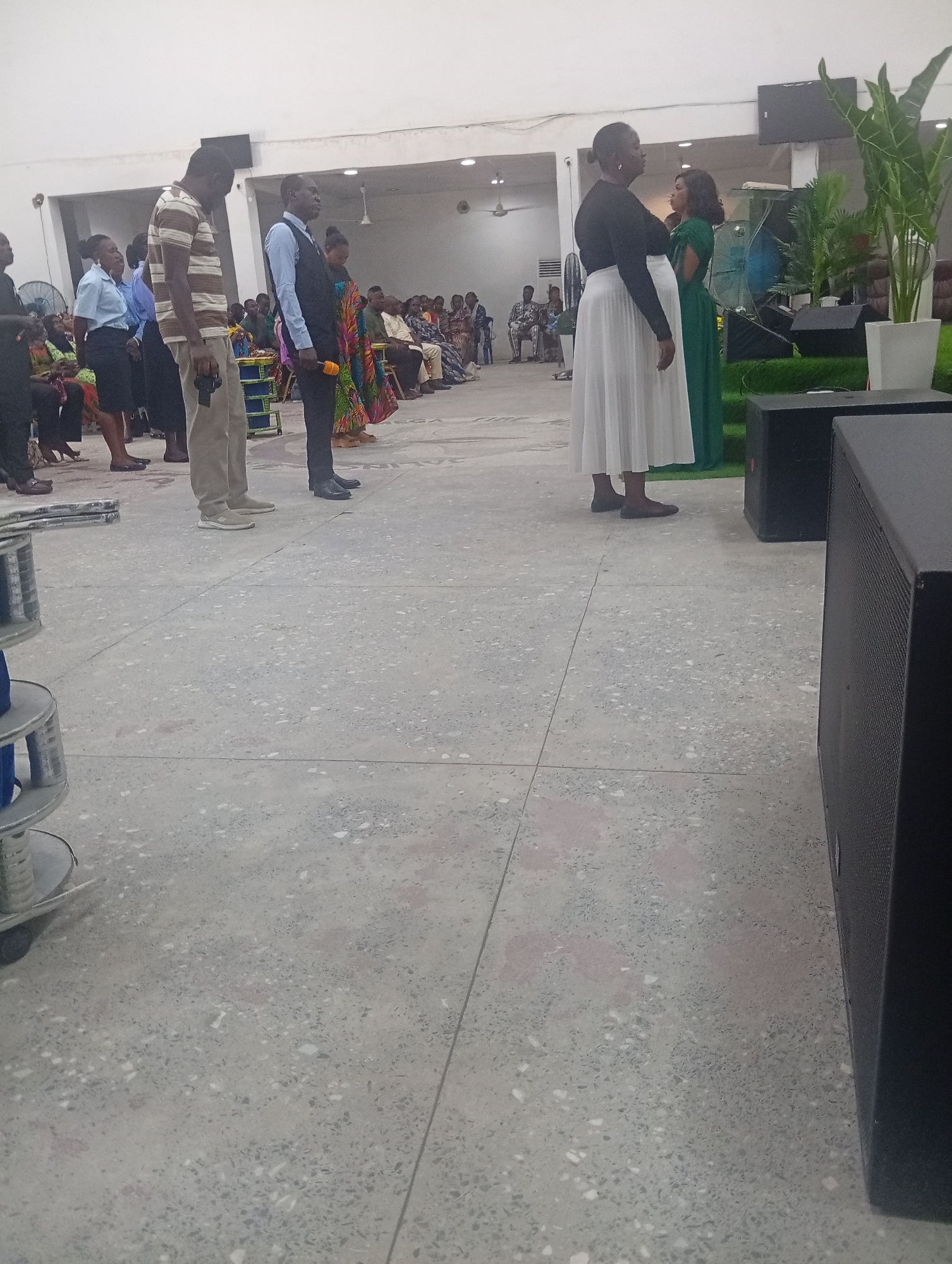 In Kason, Nigeria, a group of people stand in a line on a polished concrete floor. The group consists of at least six individuals visible from the waist down, including men and women. One man in the foreground wears a striped shirt and tan pants, holding a microphone. Next to him, another man in a dark vest and light blue shirt also holds a microphone. Further down the line, several women are dressed in traditional Nigerian attire, characterized by vibrant colors and patterns. In the background, more people are seated, and a large green plant stands in a white planter, suggesting an indoor event or ceremony. Two large black speakers are positioned in the foreground, implying a performance or public address is taking place. The lighting is bright, illuminating the scene.