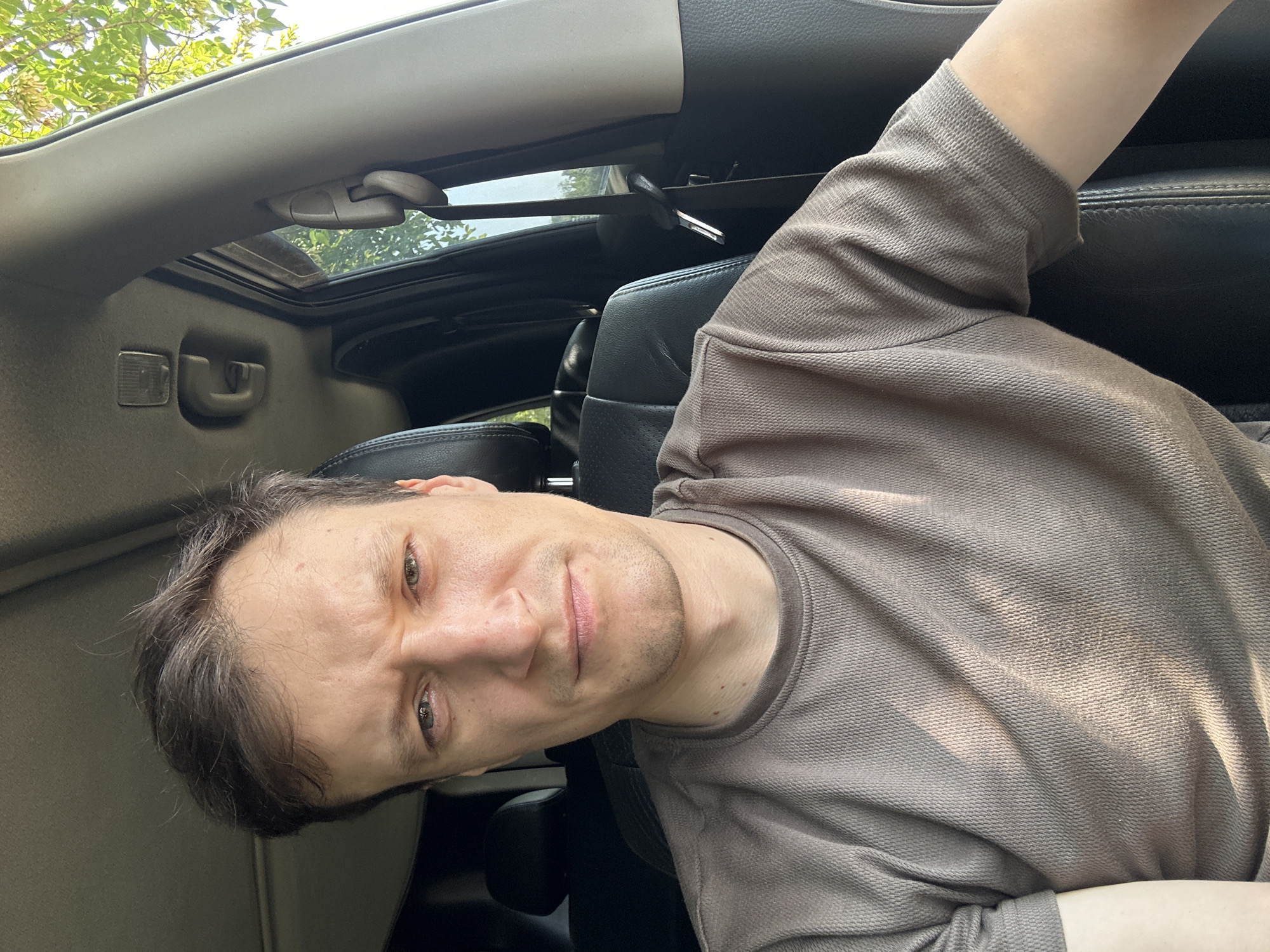 A medium shot shows a man with short brown hair and a light complexion sitting in the driver's seat of a car, looking directly at the camera. The man is wearing a brown t-shirt. Sunlight is casting shadows across his face and shirt, indicating it is daytime. The interior of the car is visible, including black leather seats and the car's ceiling. Outside the car window, green foliage suggests the vehicle is in a wooded or park-like area. There is no indication of the specific location being in Makiv, Ukraine.
