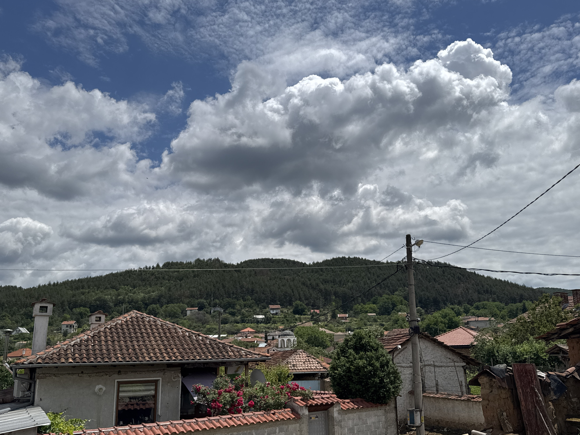 The image depicts a view of the village of Prolom, Bulgaria, on a cloudy day. The foreground features rooftops of houses with terracotta tiles, a stone wall, and some greenery, including red roses. In the midground, more houses are visible scattered across a gentle slope, interspersed with trees. A prominent feature is a forested hill that forms the background of the scene. The sky is filled with a dramatic display of large, gray and white cumulus clouds. The overall impression is of a rural, somewhat weathered village set against a natural, hilly landscape.
