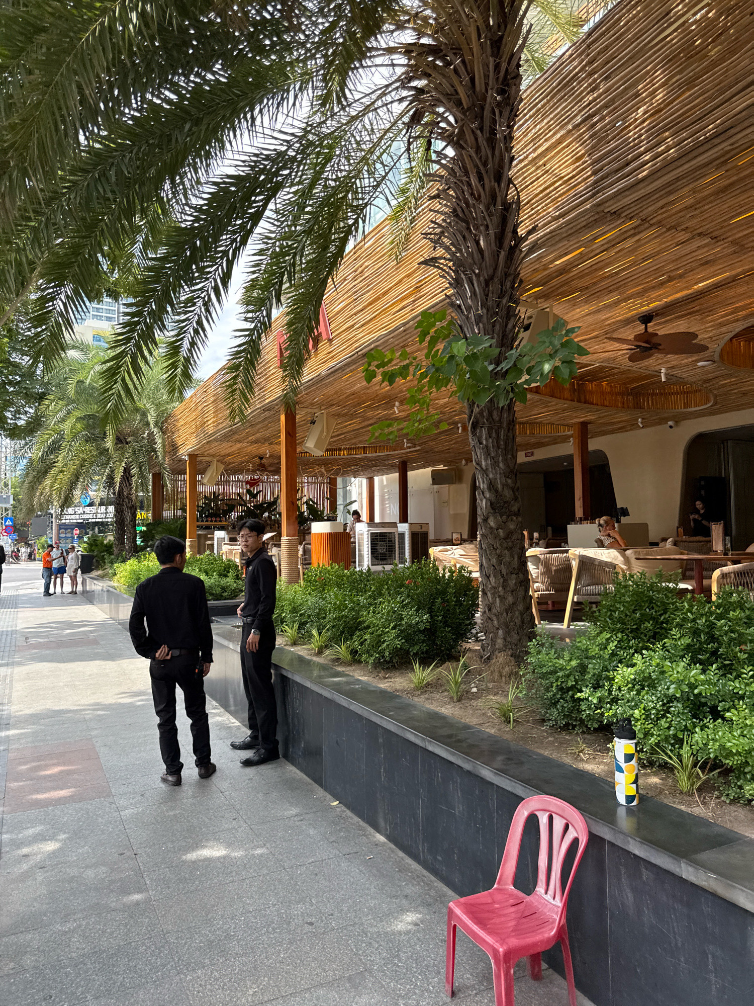 The image depicts a street scene in Nha Trang, Vietnam. In the foreground, two men in black uniforms stand on a sidewalk, facing away from the camera. To their right, a red plastic chair is placed next to a low, black planter filled with green bushes. A water bottle with a colorful, geometric pattern rests on the planter's edge. Behind the planter is a building with a thatched roof, suggesting an outdoor seating area for a restaurant or cafe. Palm trees with thin trunks and long fronds line the sidewalk, casting shadows on the ground. In the background, other people walk along the sidewalk, and a few buildings are visible. The overall atmosphere is that of a sunny day in a city.