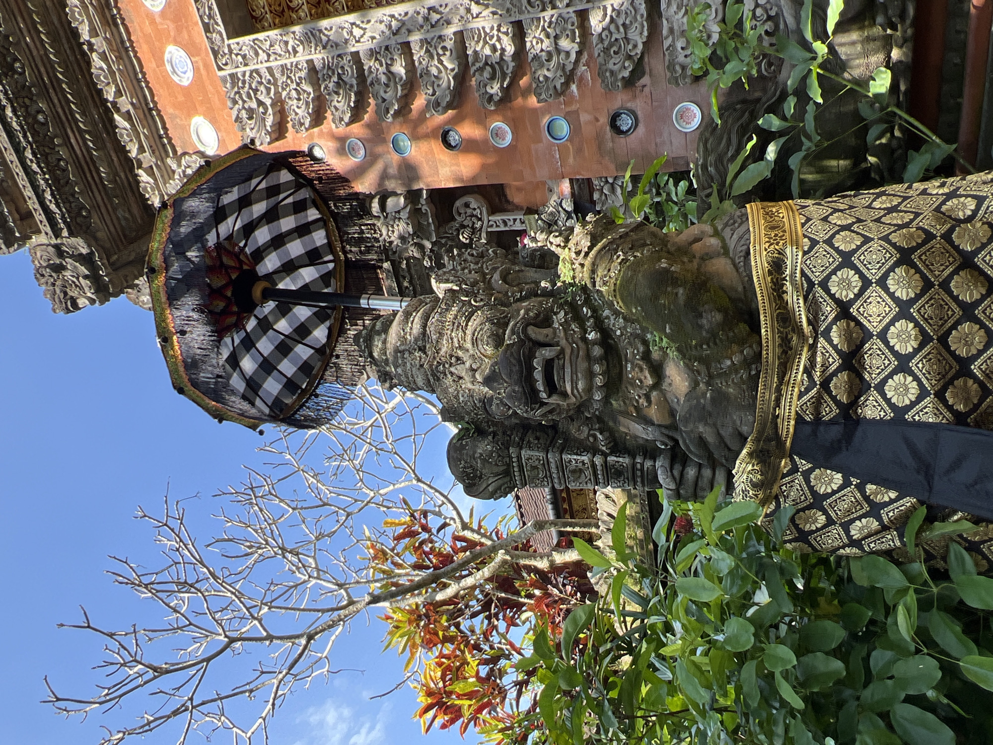 A ground-level shot in Petulu, Indonesia, angled upward toward a stone statue adorned with a black and gold patterned cloth and a black and white checkered umbrella. The statue has detailed facial features and mossy texture. A bare tree branch and lush green foliage frame the statue. Behind the statue is an ornately carved structure with a row of circular embellishments on a red background. The sky is visible in the upper left corner and is bright blue.
