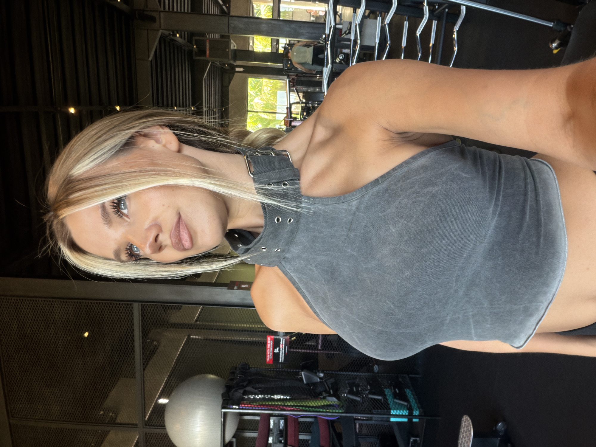 In a brightly lit gym in North Kuta, Indonesia, a young woman with blonde hair styled in a casual updo poses for a selfie. She is wearing a grey, sleeveless, high-neck crop top with metal eyelets on the collar. The background shows gym equipment, including weight racks, a stability ball, and resistance bands, along with large windows revealing a tropical outdoor environment. The woman's gaze is directed upwards and to the side, with a neutral expression. The overall impression is one of a fitness enthusiast capturing a moment in her workout routine.