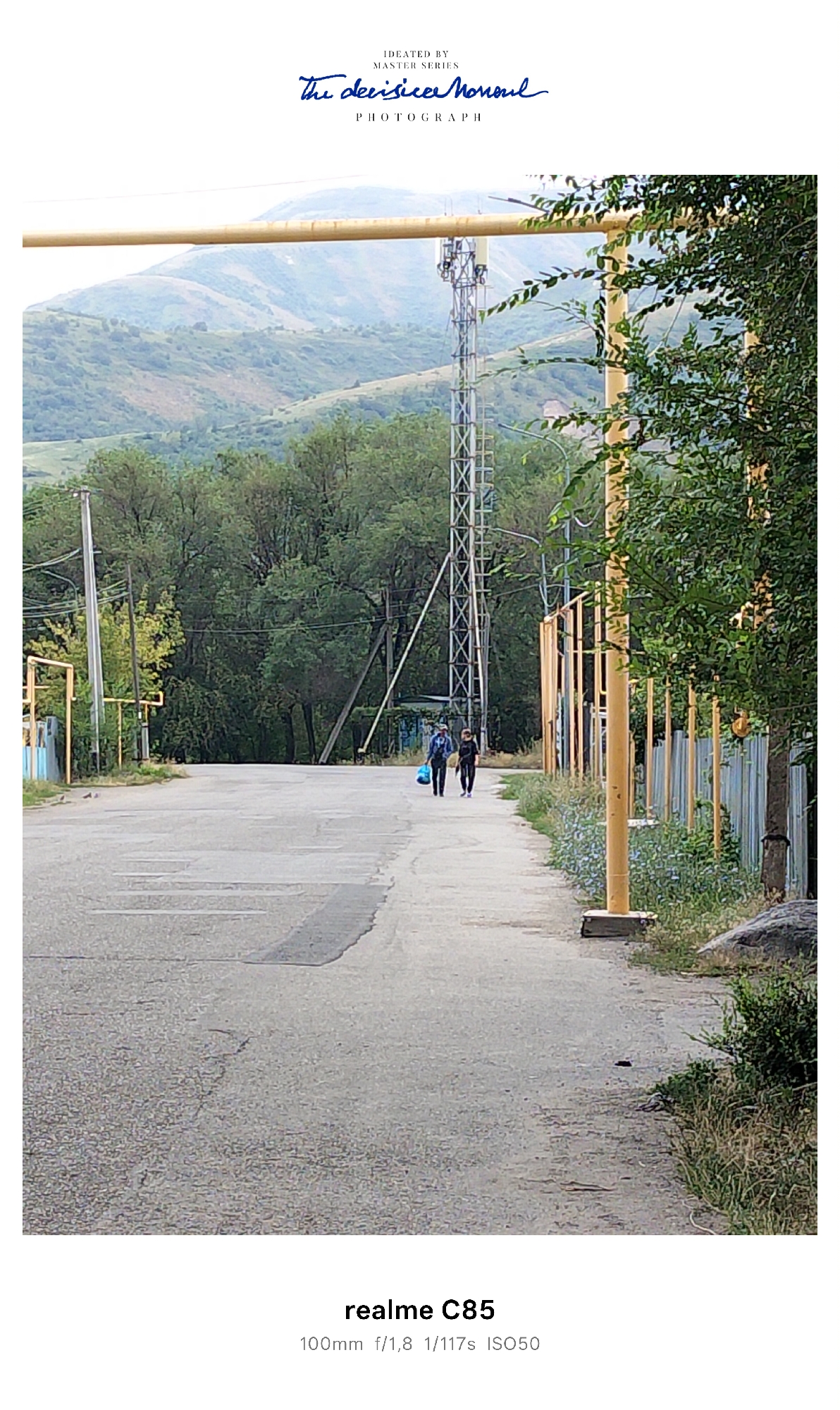 Two individuals walk down a paved road in a semi-rural or suburban area, which is identified as being in the city of Tekeli, Kazakhstan. The road appears to be asphalt with some patching and cracks. The person on the left carries a light blue bag, while the person on the right carries a dark bag. They are walking away from the viewer and towards a wooded area with hills in the background. A cell tower is visible on the left side of the road, and yellow poles and a fence line are on the right side. The environment is overcast with vegetation including trees and grass. The image also includes text overlays at the top and bottom, indicating "IDEATED BY MASTER SERIES The decisive Monoul PHOTOGRAPH" and "realme C85 100mm f/1,8 1/117s ISO50".