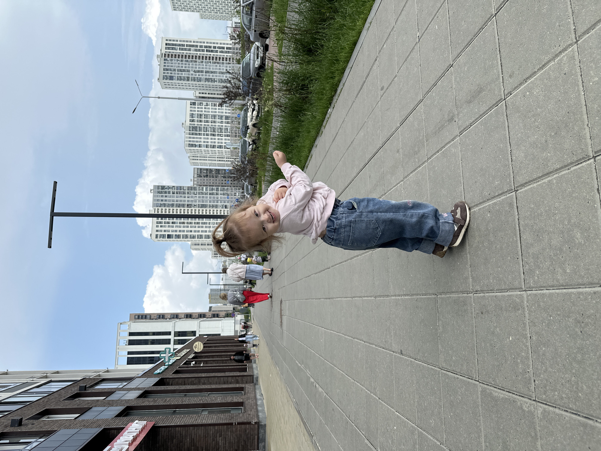 This image captures a vibrant urban scene in Yekaterinburg, Russia, featuring a young child on a sidewalk. In the foreground, slightly to the right, a young child, likely a toddler with light brown hair tied in a high ponytail, stands on a light grey paved sidewalk. The child is looking back over their left shoulder towards the camera, smiling broadly with their mouth open, revealing teeth. They are dressed in a light pink long-sleeved top, dark blue denim jeans, and brown sneakers with white soles. Their arms are bent at the elbows, with hands held near their chest.
The sidewalk, constructed from large, rectangular light grey paving slabs, extends from the immediate foreground into the distance on the left side of the frame. To the immediate right of the sidewalk, a strip of unkempt green grass and weeds separates the pedestrian area from a paved road where several cars are parked, including a white SUV, a dark sedan, and a light-colored sedan.
Along the left side of the sidewalk, a multi-story building with a dark brick facade and lighter-colored panels features multiple windows and ground-level commercial establishments. A red sign with white lettering partially visible reads "МАТЕМАТ" (likely "Магнит," a Russian supermarket chain). Further along, another storefront displays a green cross sign and text that includes "Здоровое питание" (Healthy nutrition), indicating a pharmacy and healthy food store. In the distance on the sidewalk, several adults are walking away from the camera. One woman wears a grey top and red pants, accompanied by another woman in a light-colored top and light blue skirt. A small child in yellow clothing is visible near them, and other figures are further down the path.
The background is dominated by a series of tall, modern residential buildings with numerous windows and light-colored facades, characteristic of new urban developments. Multiple black streetlights, some with single arms and others with two, punctuate the scene. The sky above is partly cloudy, exhibiting patches of bright blue interspersed with white cumulus clouds. The overall lighting suggests it is daytime, providing clear visibility of the urban environment.