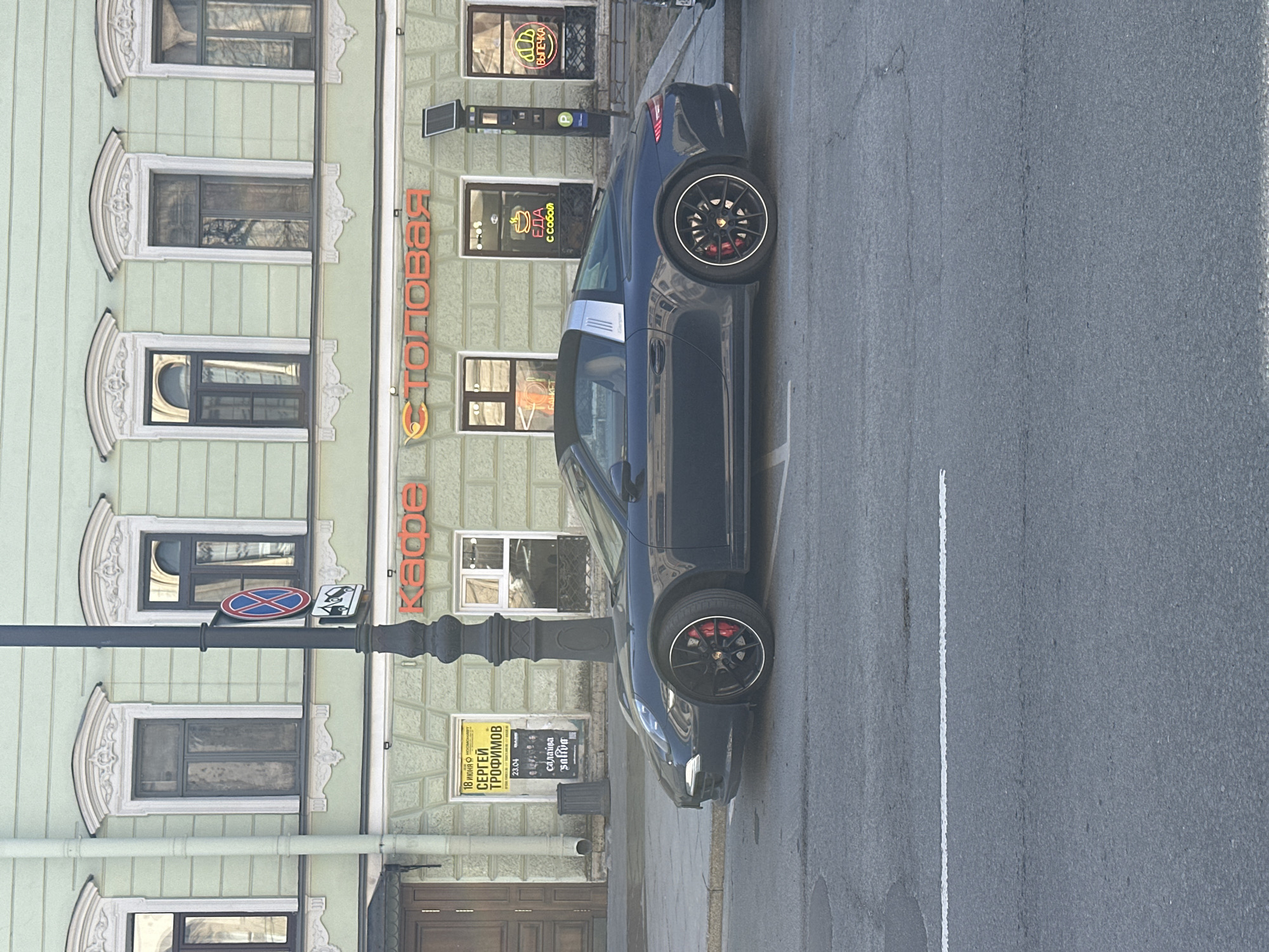 An upright-oriented shot captures a dark blue or black sports car, likely a Porsche 911 convertible, parked on an asphalt city street in Saint Petersburg, Russia. The car features black multi-spoke wheels with red brake calipers, and the Porsche crest is visible on the wheel cap. A white lane marking runs along the right side of the street.
The car is positioned alongside a light green, multi-story building with ornate white architectural detailing around its numerous windows. The ground floor of the building houses several commercial establishments. Above the car, visible signs include "СТОЛОВАЯ" (Stolovaya - cafeteria/dining hall) with a fork and spoon logo, "Кафе" (Kafe - cafe), "ЕДА с собой" (Food to go/Takeaway food) with a bowl logo, and "ВЫПЕЧКА" (Baked goods/Pastries) with a pretzel-like logo.
A black lamppost stands between the car and the building, adorned with a red circular "no stopping" sign and another blue square sign featuring a white "P" and a money symbol, indicating paid parking. On the building facade, a yellow poster advertises "18 июня космонавт СЕРГЕЙ ТРОФИМОВ" (June 18 cosmonaut SERGEY TROFIMOV) with "23.04" and "салайва saliva" also printed on it. The time of day appears to be daytime, possibly late morning or afternoon, with soft, indirect lighting. No people are visible in the scene.