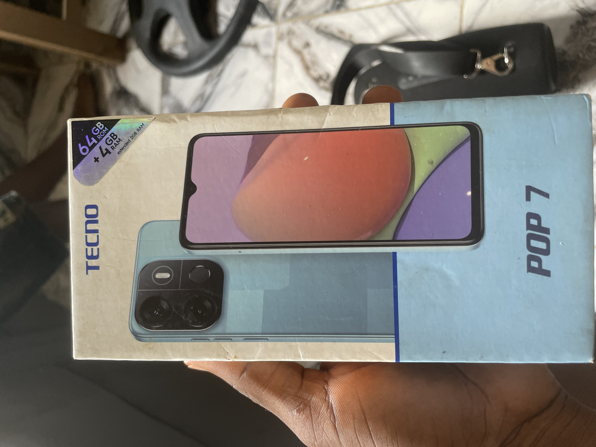 A light blue cardboard box for a TECNO POP 7 smartphone is being held by a person's left hand. The box features a picture of the phone on the front, showing its light blue back with a dual-camera setup and a purple and orange-toned screen. A sticker in the upper right corner of the box indicates "64GB ROM + 4GB RAM extended 2GB RAM". The background is out of focus but appears to be an indoor setting with tiled flooring and furniture. The person's hand is dark-skinned and partially visible on the left side of the image. The scene is likely set in Keffi, Nigeria, given the context of the product.
