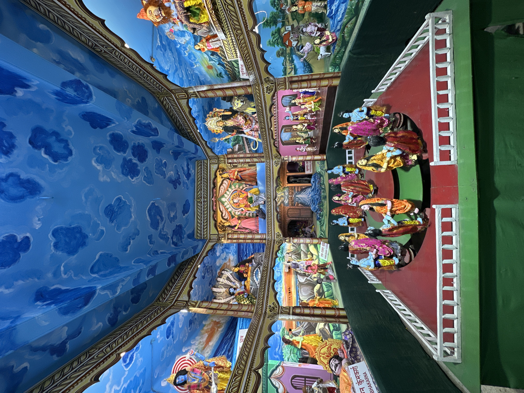 This is a close-up, indoor shot taken from below, focusing on a series of traditional Hindu dioramas and paintings.
The ceiling is painted with a light blue sky and white clouds. Below this, there are several framed dioramas depicting scenes from Hindu mythology. These include depictions of:
* Narasimha
* Durga riding a lion.
* Rama-Ravana Yuddha (Rama-Ravana War)
* Krishna-Janm
Each diorama is adorned with intricate details and vivid colors. At the bottom, there is a structure with a red-painted surface and a white staircase, where several figures, likely representations of deities or characters from Hindu stories, are arranged.
A label in Hindi is visible near the bottom right corner, possibly providing information about one of the depicted scenes or figures.
The location appears to be an indoor exhibit or display, possibly in a religious or cultural center, in Varanasi, India.