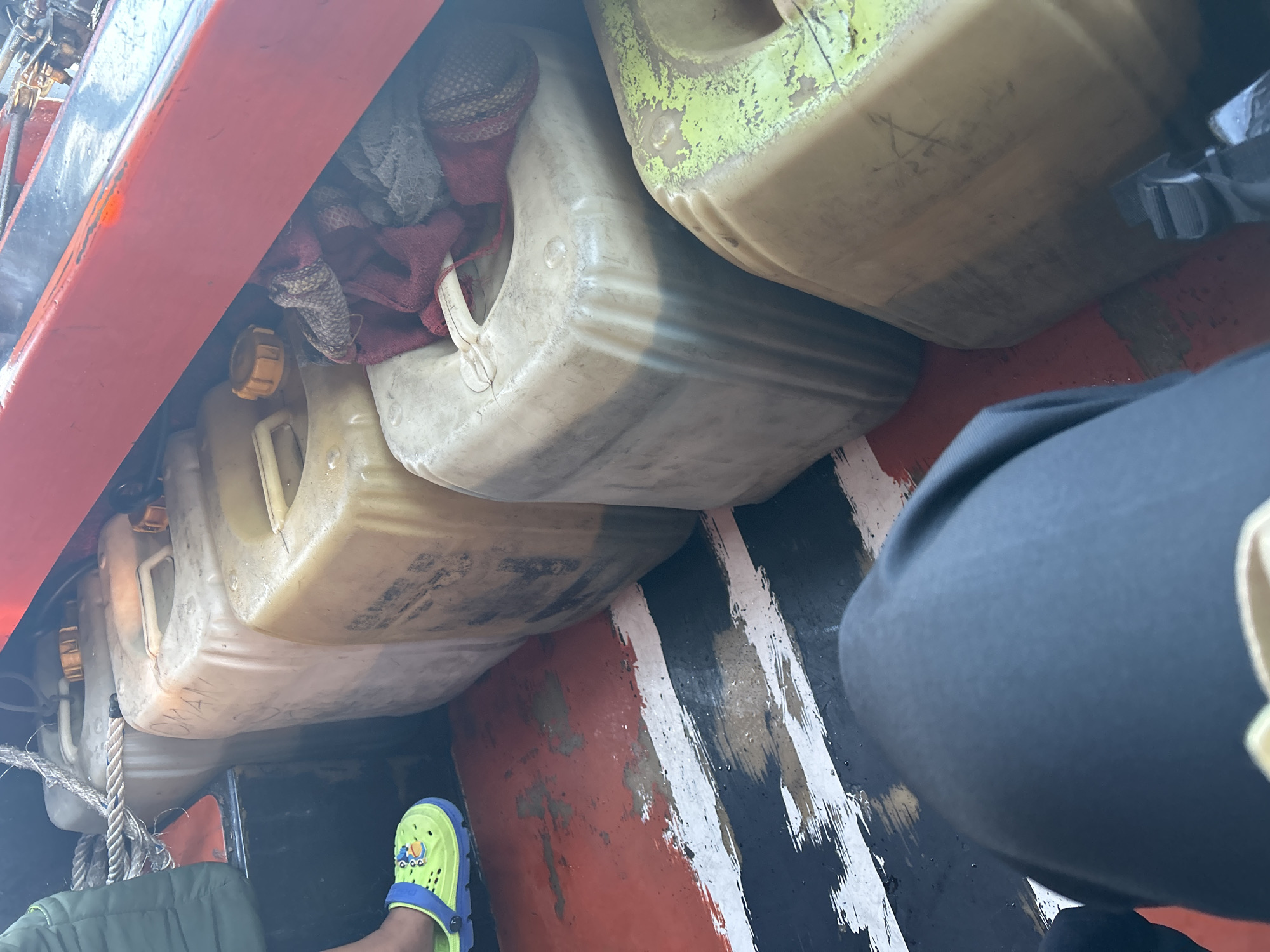 In an unknown location in Indonesia, multiple plastic jerrycans, some filled with a dark liquid, are stored in the back of a vehicle, possibly a boat. A person's legs are visible in the foreground, one wearing a lime green clog with cartoon characters. Ropes are securing the jerrycans. The floor of the vehicle has a painted pattern of red, white, and black stripes. A red railing is visible above the jerrycans.