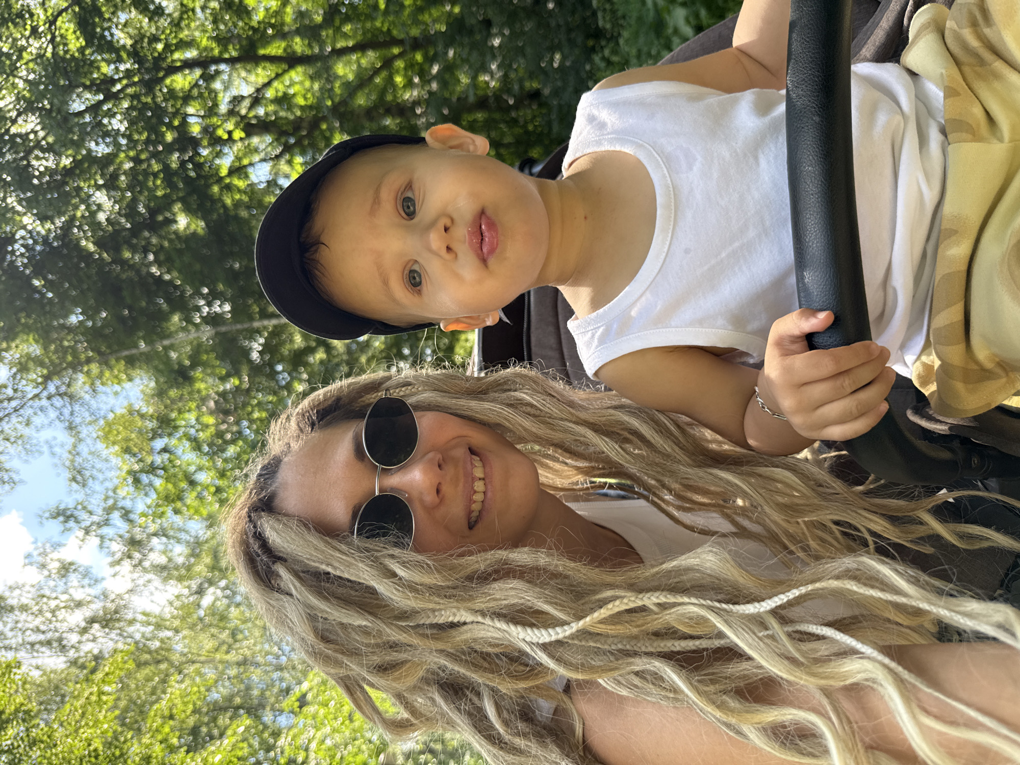 In Rusanovo, Russia, a woman with long, blonde dreadlocks and sunglasses is captured in a selfie with a young child. The woman is wearing a white tank top, and the child is wearing a white tank top and a black baseball cap. They are both outdoors, with lush green trees and a blue sky visible in the background. The child appears to be sitting in a stroller, with the woman partially obscured by the stroller's handle. The overall scene suggests a casual outing on a sunny day.