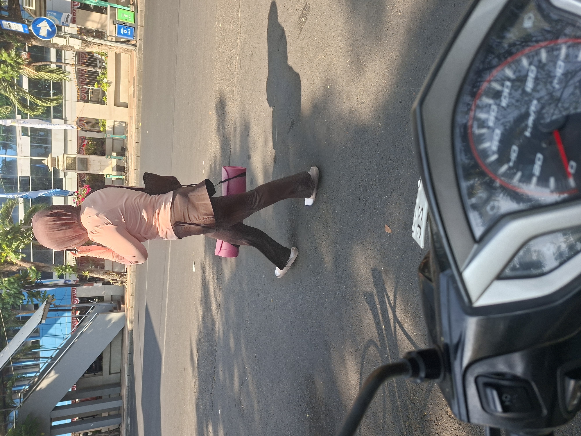 A person wearing a hijab and a light pink shirt with brown pants is walking across a street in Surabaya, Indonesia. The person is carrying a rolled-up pink yoga mat under their right arm and has a black bag slung over their left shoulder. They are wearing white sandals. In the foreground, the handlebars and dashboard of a motorcycle are visible. The background shows a pedestrian bridge, trees, buildings, traffic signs, and two trash cans. The sun is casting shadows on the asphalt road.