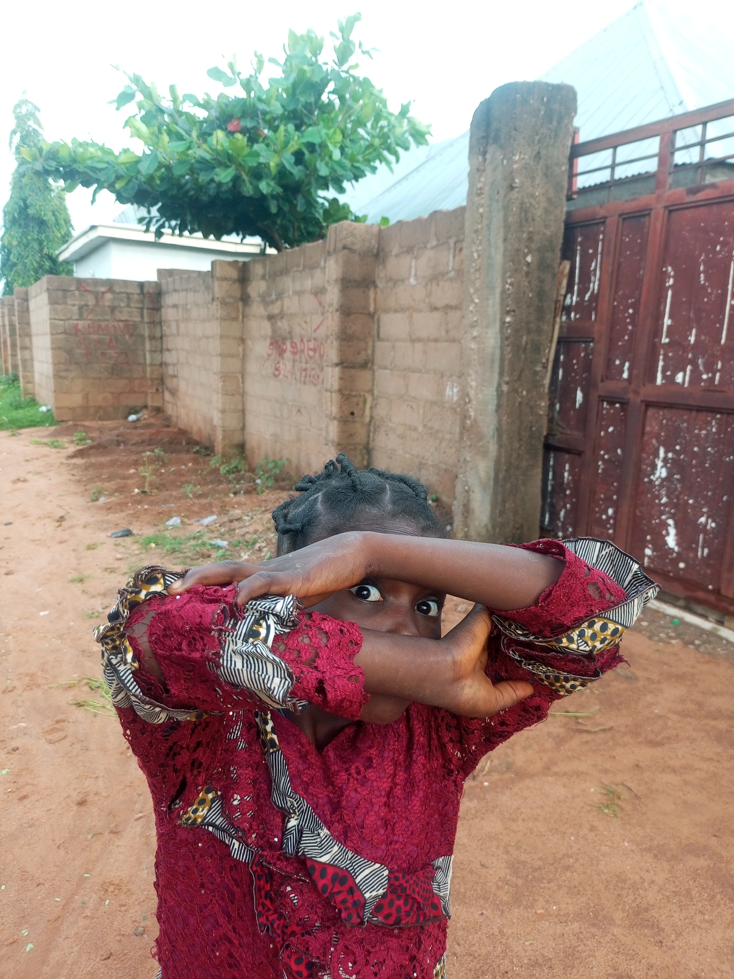 In a dusty, outdoor setting that appears to be the city of Tempure, Nigeria, a young child with dark skin and braided hair is captured in a medium shot. The child wears a vibrant, textured crimson dress with intricate lace detailing and bold patterned fabric inserts. Their arms are raised, and their hands are positioned in front of their face, partially obscuring it and leaving only their wide, dark eyes visible and looking directly at the camera.
The background is composed of a rough, brick wall constructed from lighter colored, irregular bricks, some of which are marked with faded red graffiti. To the left of the wall, a tall, slender tree with lush green foliage stands, and beyond that, a portion of a light-colored building with a steep, metallic roof is visible. To the right of the brick wall is a tall, rough-hewn concrete pillar, and behind that, a tall, reddish-brown metal gate, partially ajar, revealing a glimpse of a corrugated metal roof behind it. The ground is a mixture of reddish-brown dirt, scattered with small clumps of dry grass and some white debris, suggesting an unpaved area. The overall lighting suggests an overcast day.
