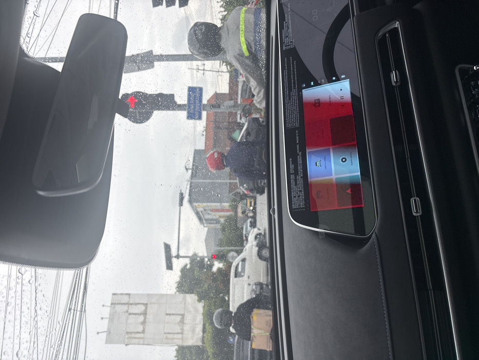 A car interior view shows a digital dashboard display featuring red and blue square icons. Through the rain-streaked windshield, a street scene includes a red traffic light, utility poles, and various pedestrians and motorcyclists, some wearing helmets. A white van is also visible. The rearview mirror is positioned above, reflecting the street scene.