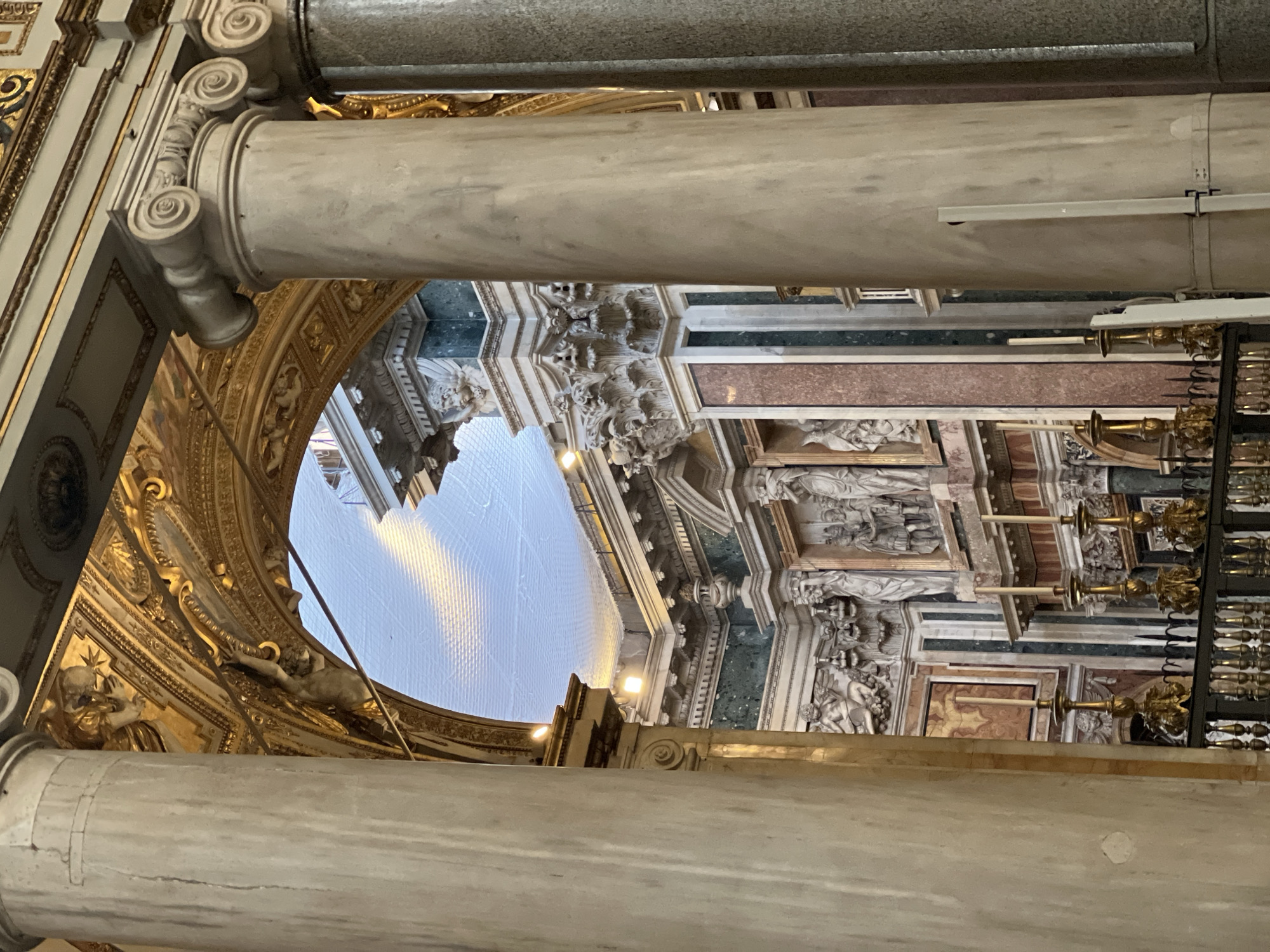 This image captures the opulent interior architecture of the Basilica Papale di Santa Maria Maggiore in Rome, Italy. The foreground is framed by two substantial marble columns: a light beige-grey column on the left and a similar column with an Ionic capital on the right, partially obscuring a darker column behind it. Centrally, a large gilded archway, richly adorned with intricate gold relief work and sculpted figures, spans between the columns; one figure appears to be dynamically suspended from the upper left interior curve of the arch. Through this arch, a further section of the basilica is visible, featuring a vaulted ceiling covered by a light-colored protective netting, illuminated by several artificial spotlights. The background architecture displays walls paneled with polished dark green and reddish marble, contrasting with white marble elements and elaborate white stucco or carved stone decorations. Numerous classical sculptures are integrated into these walls, including winged putti, and larger figures standing within niches. At the bottom, a dark metal balustrade supports multiple ornate golden candelabras, each holding a white candle. No individuals are present in the scene.