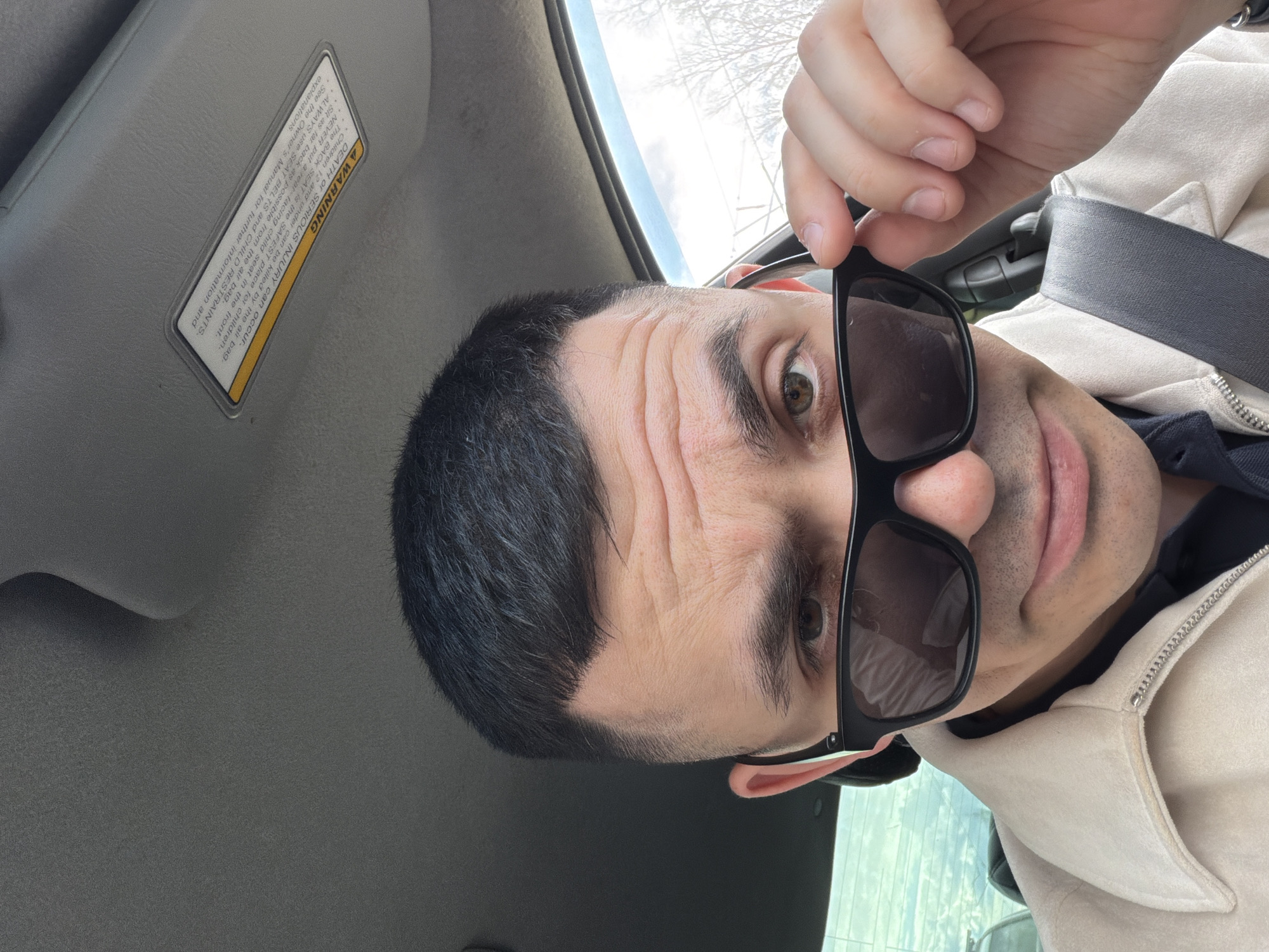 A man takes a close-up selfie inside a vehicle, likely in Izhevsk, Russia. He is positioned towards the right of the frame, looking directly into the camera. The man has short, dark hair and is wearing a light-colored jacket over a dark shirt, with a gray seatbelt visible across his chest. His right hand holds dark-framed sunglasses, slightly lowered to reveal his right eye, which appears light brown or hazel. His face shows several horizontal wrinkles on his forehead, and his expression is neutral with slightly parted lips.
The car's interior is visible, featuring a gray ceiling and a sun visor on the left side of the frame. Through the car window in the upper left, a bright sky and bare tree branches are visible, suggesting daytime during winter or early spring.
A prominent warning label on the sun visor provides safety information, stating: "A WARNING DEATH OR SERIOUS INJURY can occur. Children 12 and under can be killed by the air bag. The BACK SEAT is the SAFEST place for children. NEVER put a rear-facing child seat in the front. Sit as far back as possible from the air bag. ALWAYS use SEAT BELTS and CHILD RESTRAINTS. See the Owner's Manual for further information and explanations." The man is the sole subject, engaged in taking a self-portrait while seated in the car.