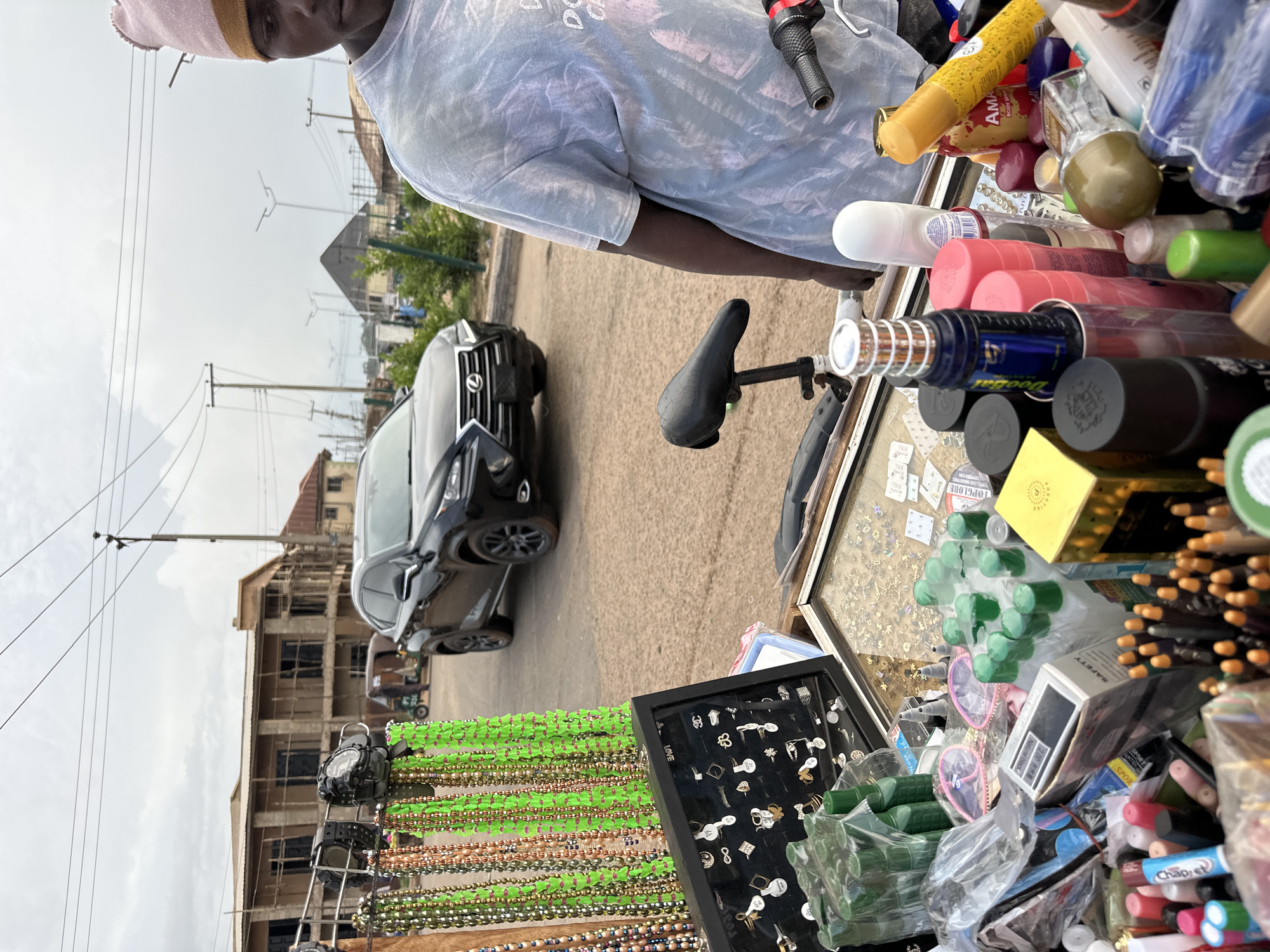 This is a medium shot, eye-level photograph of an outdoor market stall in Bukuru, Nigeria. A young Black male, wearing a light blue tie-dye t-shirt and a pink and beige beanie, stands to the right, partially obscuring the view of the stall's contents. The stall itself is filled with various items, including strings of colorful beads, rings displayed in a black case, bottles of toiletries, and other assorted products. In the background, a black Lexus SUV is parked on a dirt road. Further back, unfinished buildings and power lines are visible, suggesting a developing urban area. The sky is overcast. The overall impression is of a typical street vendor scene in a Nigerian city.