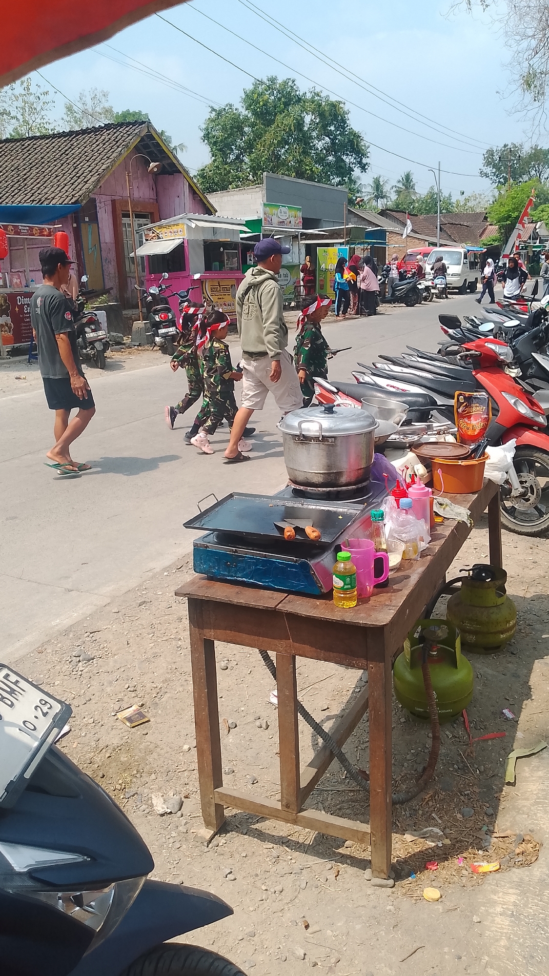 This image depicts a street scene in Karanglangu, Indonesia, likely during a sunny day. In the foreground, a wooden table is set up as a food stall. On the table are cooking utensils, including a large pot, a grill with food items, several bottles of condiments and drinks, and two green propane gas cylinders connected by hoses to the cooking equipment.
Behind the food stall, a group of children in camouflage uniforms with red and white headbands are walking in procession. They appear to be participating in a parade or event. Two adults are accompanying them, one in a light-colored hoodie and shorts, and another man in a dark t-shirt and shorts. The man in the dark t-shirt is on a small skateboard-like device.
The background shows a street with several parked motorcycles and a few buildings, including small shops or stalls with colorful facades and signage. Further in the background, there are houses with tiled roofs and trees. The overall environment suggests a lively, possibly festive, atmosphere in a semi-urban or rural Indonesian setting.