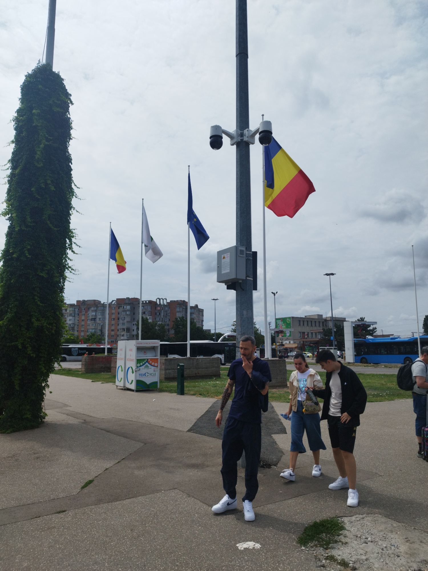 The image depicts an outdoor scene in Constanța, Romania. In the foreground, three individuals are standing on a paved walkway. The person on the left, a man, is wearing a dark blue t-shirt and dark pants, with tattoos visible on his left arm. He appears to be smoking. To his right, a woman is wearing a light-colored jacket over a white top and denim culottes. Next to her, a young man is dressed in a white t-shirt, black shorts, and a black jacket.
Dominating the center of the image is a tall metal pole with two security cameras mounted on it. A Romanian flag, red, yellow, and blue, is flying from a flagpole attached to the same pole. Behind the people and the pole, a street is visible with several buses, including a blue one, and buildings in the background. One building appears to be a block of apartments. There are also a few flags flying from poles further back, one of which is a blue European Union flag. To the left of the main pole, a tall structure covered in dense green ivy stands out. In front of the buildings, a white recycling bin with the inscription "TEXCYCLE" is visible. The ground is paved with concrete slabs and there are patches of grass. The sky is overcast with clouds.