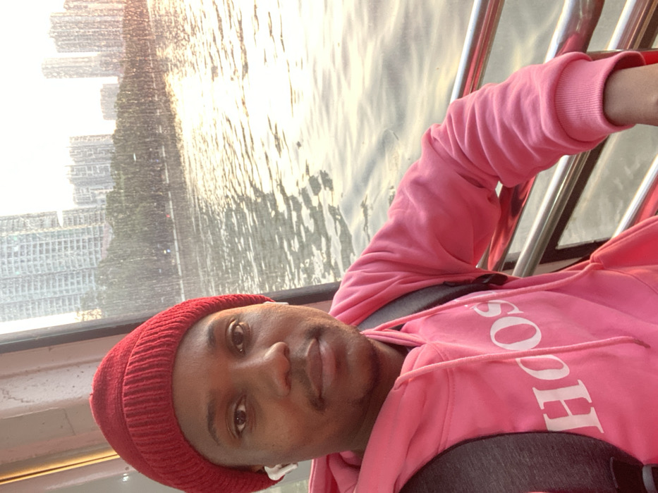 The image shows a close-up portrait of a young adult, likely male, aboard what appears to be a boat or ferry. He has dark skin, is wearing a bright red knitted beanie, and a vibrant pink hooded sweatshirt. White wireless earbuds are visible in his ears. He looks directly at the camera with a neutral expression. Dark backpack straps are visible over his shoulders.
The background features a wide body of water, possibly a river or bay, brilliantly reflecting golden, warm light, suggesting either sunrise or sunset. The water's surface is covered in ripples. Beyond the water, a dense and modern cityscape rises, characterized by numerous tall buildings forming a prominent skyline. Some greenery and what appears to be a road are visible along the distant bank. Silver metal railings, indicative of a boat's structure, are present to the right of the person.
The overall atmosphere is illuminated by the soft, glowing light of dawn or dusk. Part of a white logo, spelling "SOOH", is visible on the front of his pink hoodie. This scene is set in Guang Zhou Shi, China, depicting a moment on the water with the city as a backdrop.