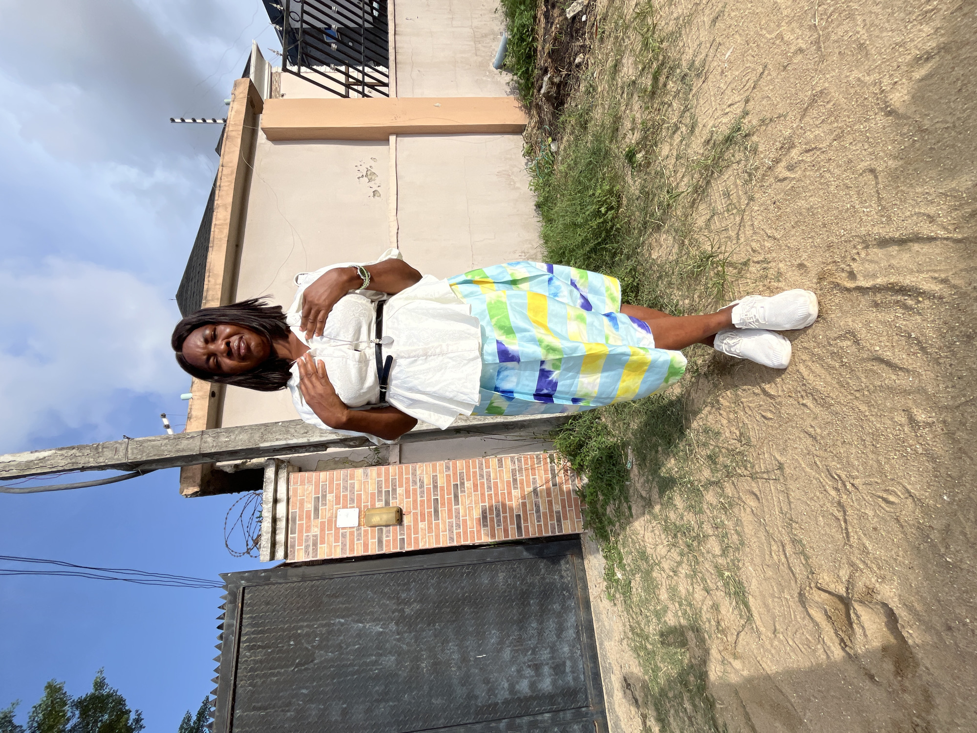 A single adult female is depicted standing outdoors on a sandy patch of ground in Iranla, Nigeria. She faces the viewer directly, with her eyes partially closed and her mouth slightly downturned. She wears a white, textured short-sleeved blouse, a thin black belt, a long silver-colored necklace with a dark pendant, a multi-colored striped skirt (blue, light green, yellow, white), and white athletic shoes. Her hands are positioned over her upper chest/abdomen.
The background consists of a residential street scene. To the immediate left is a tall, dark metal gate with a diamond plate pattern, adjacent to a brick pillar topped with coiled barbed wire. A light-colored electrical box and a brass-colored light fixture are mounted on this pillar. A concrete utility pole with overhead electrical wires stands centrally in the mid-ground. Behind the subject and to the right are various residential buildings with light-colored, possibly beige, exterior walls and dark roofs. One building's roofline shows a striped black and white pole. The ground is dry earth with sparse green grass patches. The sky is partly cloudy with areas of clear blue visible.