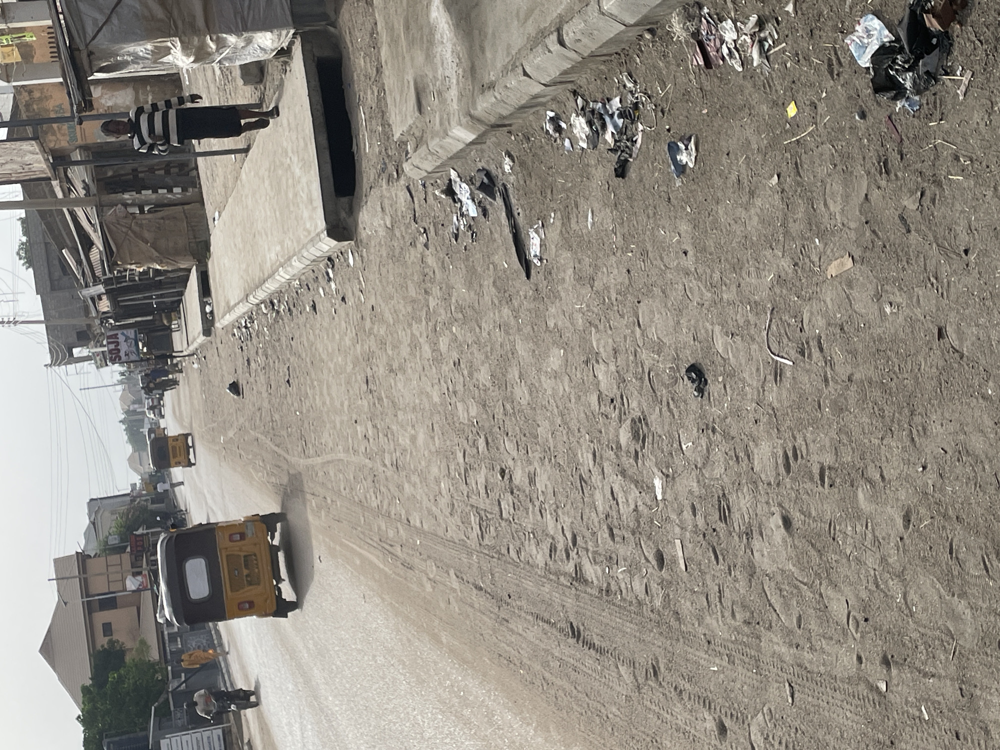 The image depicts a street scene in Unknown, Unknown. The street is mostly barren, with patches of litter on the side. A yellow tricycle is moving along the street. A person wearing a striped shirt is walking along the sidewalk. Several buildings line the street. The environment appears to be dusty.