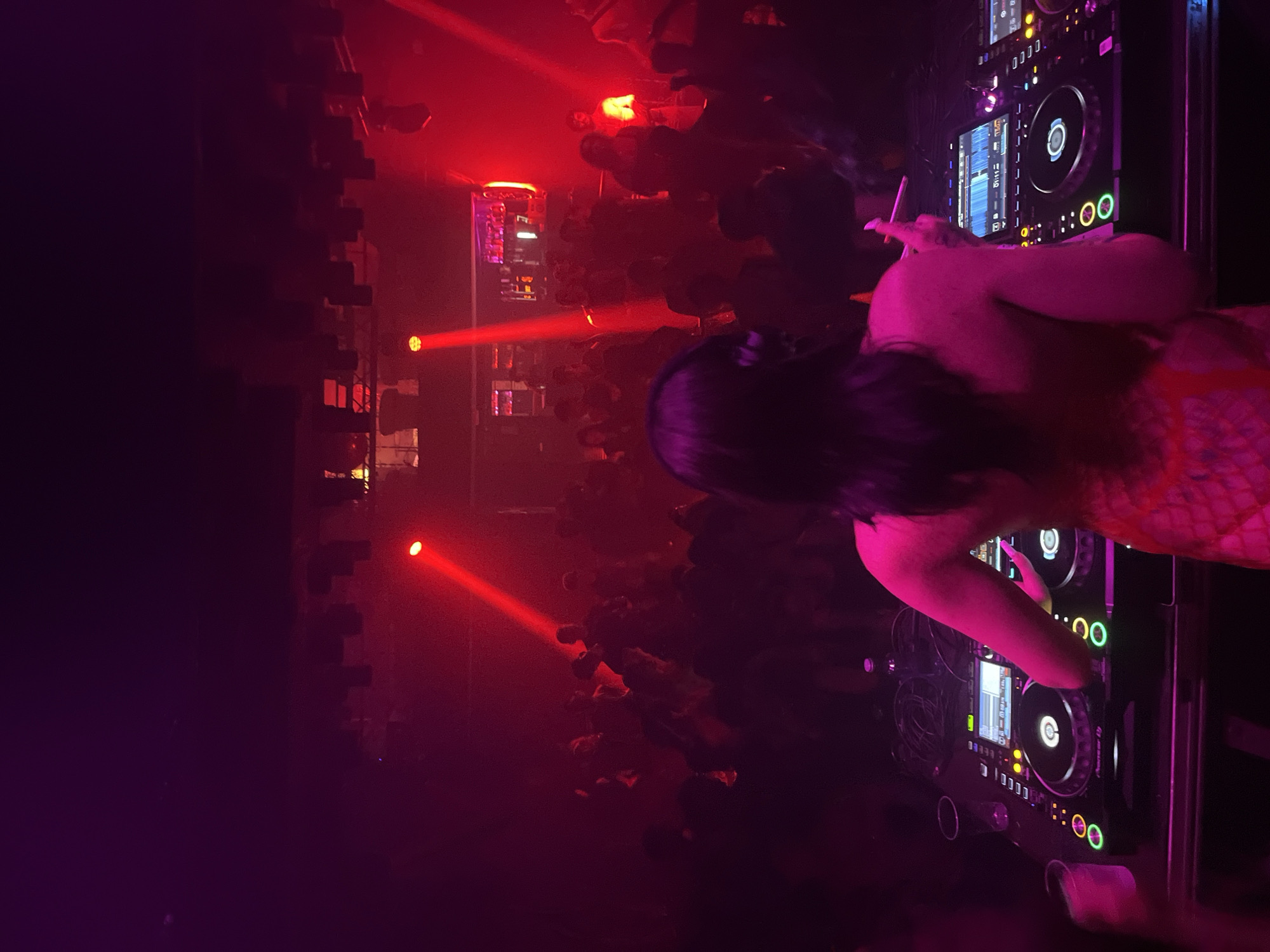 A DJ performs in a nightclub. The nightclub is dimly lit in red and purple lighting. A crowd of people is visible on the dance floor. The DJ has long dark hair. They are wearing a patterned top. The DJ is holding a cigarette in one hand and manipulating the controls on a professional DJ mixer in the other. The mixer includes a pair of turntables with screens. The nightclub may be in Barcelona, Spain.