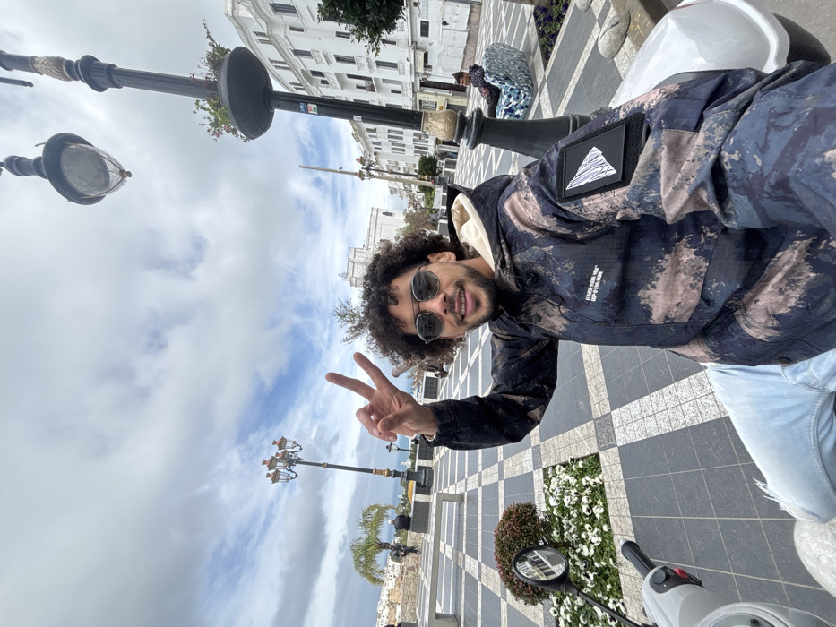 A male individual is depicted in a medium shot, smiling directly at the camera and making a "peace sign" with his right hand. He possesses dark, curly hair and a beard, and wears round sunglasses. His attire consists of a hooded jacket featuring a distressed camouflage or abstract pattern in shades of navy blue, light blue, and tan, with a visible light-colored hood lining and a black patch with a white mountain motif on the right sleeve. He is also wearing light blue ripped jeans and white shoes. The subject is seated on a light grey/white scooter, with its handlebars and rearview mirror visible in the lower left quadrant.
The scene takes place outdoors in **Place du 9 Avril 1947 (Grand Socco) in Tangier, Morocco**. The plaza floor is paved with a distinctive black and white checkered tile pattern, interrupted by planted garden beds containing white flowers and green foliage. In the mid-ground, an ornate dark streetlamp with gold accents and an attached flower planter is positioned to the right. A second similar streetlamp is visible further back on the left. A large, dark cannon is situated in the mid-background, left of the central subject. The background features several white, multi-story buildings exhibiting traditional Moroccan architecture. A prominent white historic building with multiple arched windows is visible in the far background. The sky above is partially cloudy, revealing patches of blue. One male is observed seated on a distinctive, colorful patterned bench in the mid-ground to the right. Several other indistinct figures are present in the distant background.