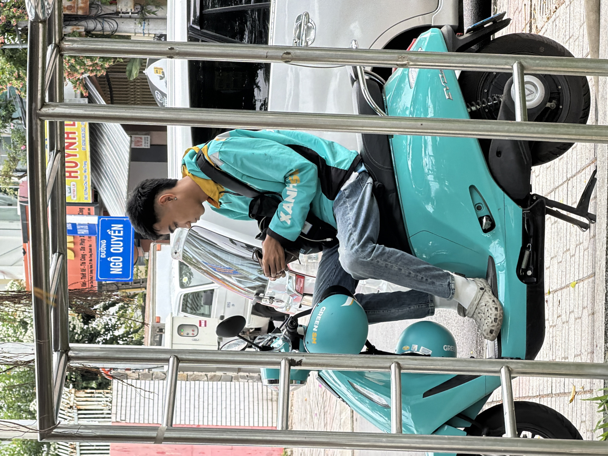 A young Vietnamese male, identified as Vietnamese, is seated on a turquoise electric scooter, identified as an Eviny, on a city street in Nha Trang, Vietnam. He is wearing a teal and yellow hoodie with black accents, blue jeans, white socks, and gray Crocs. He is holding a black smartphone in his hands and looking down at it. A teal helmet with the words "GREEN SM" is hanging from the scooter's handlebars. The scooter is parked under a metal canopy structure. In the background, there is a white van, a silver car, and various buildings, including a sign with Vietnamese text and an advertisement for "HUỲNH LAI". A blue street sign with white text reads "ĐƯỜNG NGÔ QUYỀN". The street is paved with light-colored stones. The scene appears to be an urban setting during the daytime.