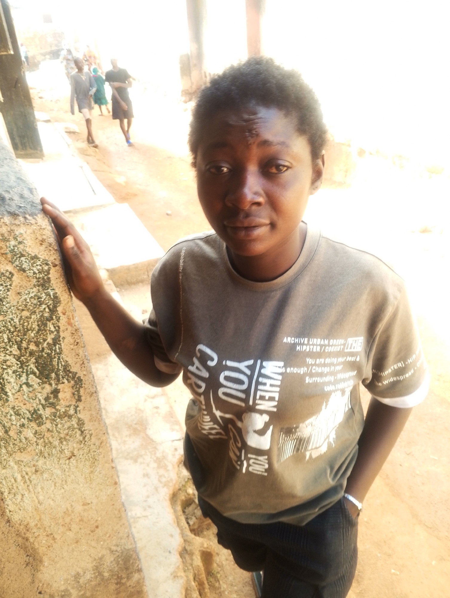 A medium shot depicts a young Black woman in a t-shirt with the words "ARCHIVE URBAN GREEN HIPSTER/COEXIST" and "WHEN YOU CART YOU" on it, standing near a weathered wall. Her hand rests on the wall, while her other hand is in her pants pocket. In the background, other people are visible walking down a dirt road. The environment appears to be a semi-urban area, possibly in Jos, Nigeria.