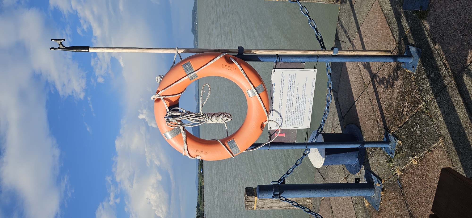 A life preserver ring hangs on a wooden pole next to a blue chain fence on a pier. The life preserver is orange with reflective silver markings and a coiled white rope. A sign is attached to the pole below the life preserver. The pier is paved with reddish-brown and gray interlocking pavers. A dark blue mooring bollard is visible behind the fence. The water is calm and reflects the sky, which is blue with scattered white clouds. In the distance, a green, hilly shoreline is visible with trees. The scene is located in Balatongyörök, Hungary.