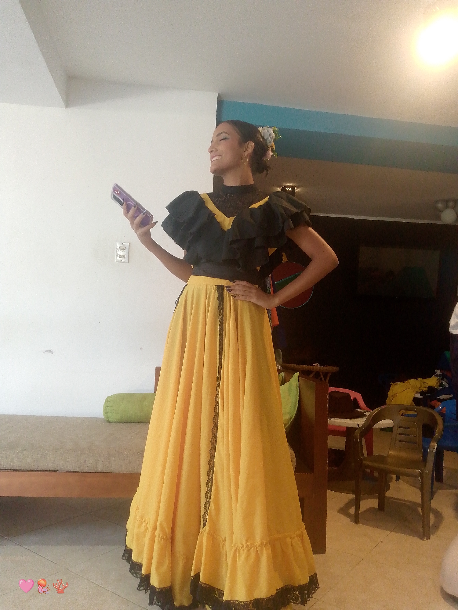 A woman stands in a room with a tiled floor, appearing to be indoors, somewhere in Playa El Agua, Venezuela. She is wearing a traditional dress consisting of a black ruffled top and a long, yellow skirt with black lace trim. She is holding a phone in her left hand and has her right hand on her hip. Her hair is styled up, with flowers. Behind her is a wall, a daybed with a green pillow, various pieces of furniture, and miscellaneous items that suggest a lived-in space. The room is generally well-lit.