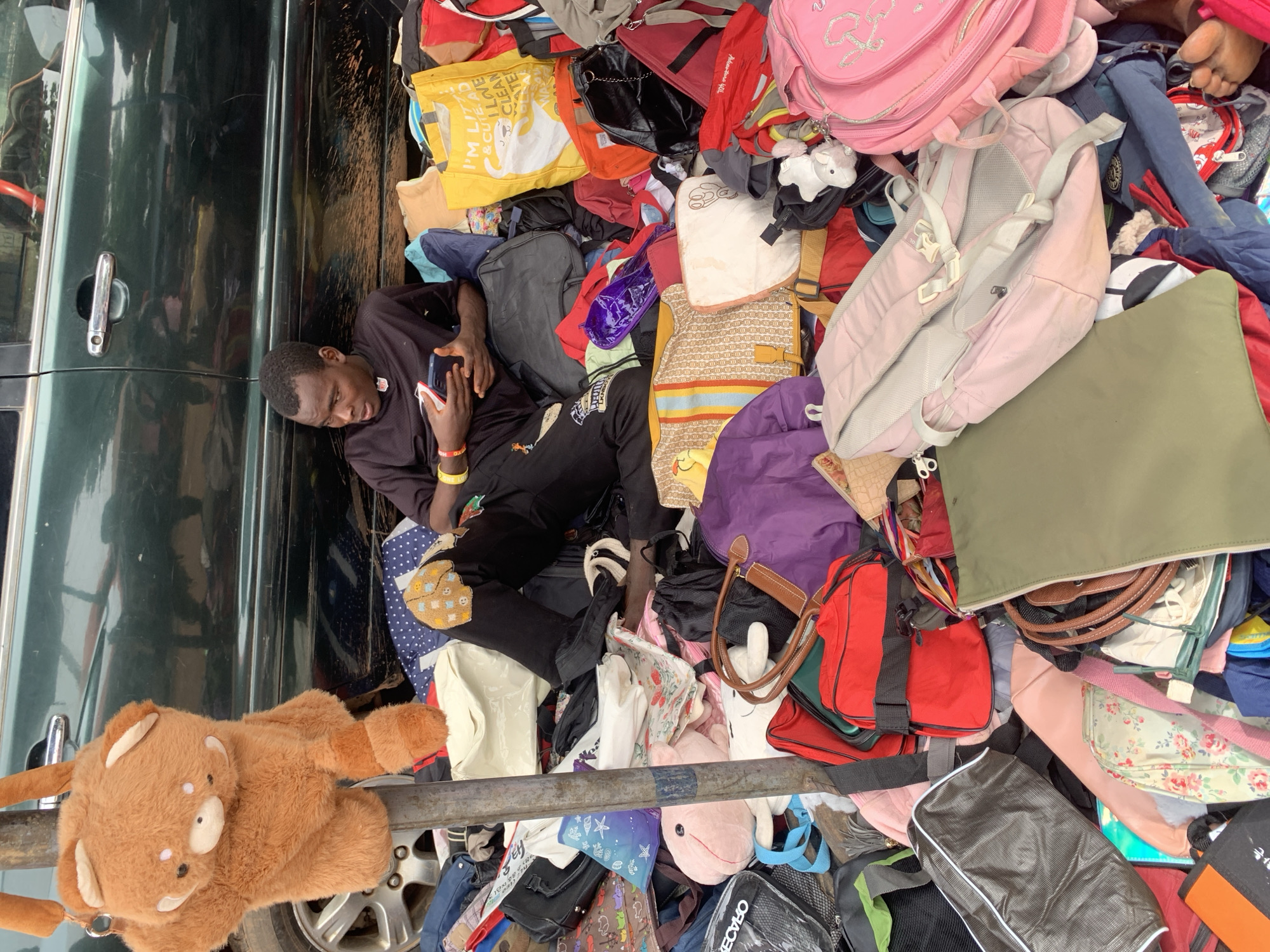 A young man is lying down amidst a large pile of bags, backpacks, and luggage. He is wearing a black t-shirt and dark pants, and is holding a smartphone in his right hand. He appears to be looking at the phone. The bags are of various colors and sizes, including pink, yellow, purple, red, green, and black. Some are brightly colored, while others are more subdued. There are also some stuffed animals visible in the pile, including a teddy bear and a pink whale. The scene appears to be outdoors, possibly a street or a market, given the presence of a dark green vehicle on the left side of the frame with a chrome door handle and a car wheel visible at the bottom. The ground is covered in bags and the overall impression is one of abundance or perhaps a chaotic sale or storage situation. The lighting suggests it is daytime. There is text on a yellow bag that reads "I'M LITT & CHILD FRIENDLY".