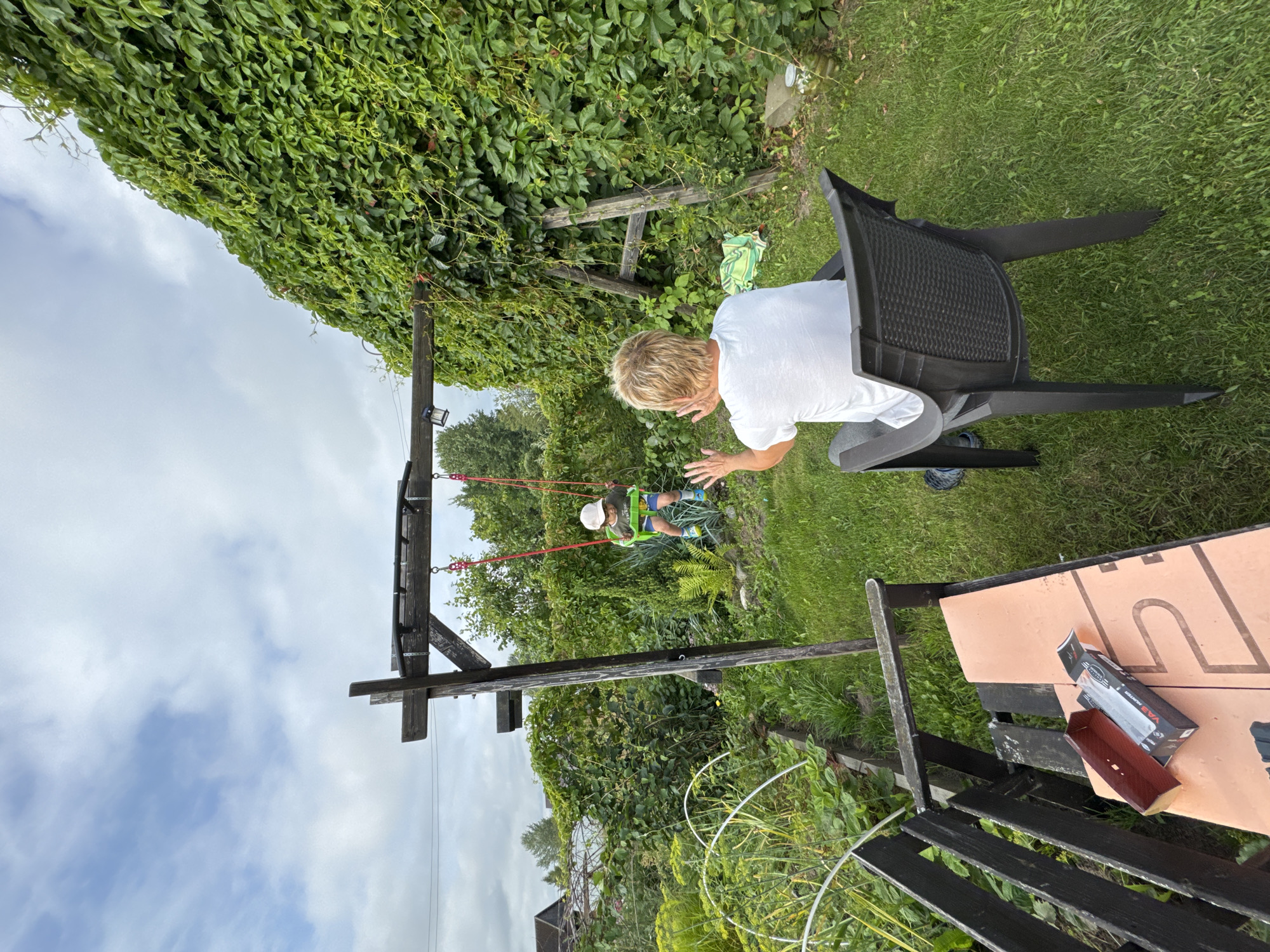 In the backyard of a property in Sitno, Russia, two individuals are present. In the background, a child sits on a swing, propelled by an unseen force, wearing a light-colored cap and blue shorts. In the foreground, an adult with blonde hair, dressed in a white t-shirt, sits in a dark plastic chair, facing away from the camera and towards the child on the swing, with their hands raised. The yard features lush green grass, a wooden swing set structure, and a dense wall of green ivy covering a wooden fence or structure. Partially visible in the lower left is a wooden bench with a box and packaging on it. The sky above is partly cloudy.