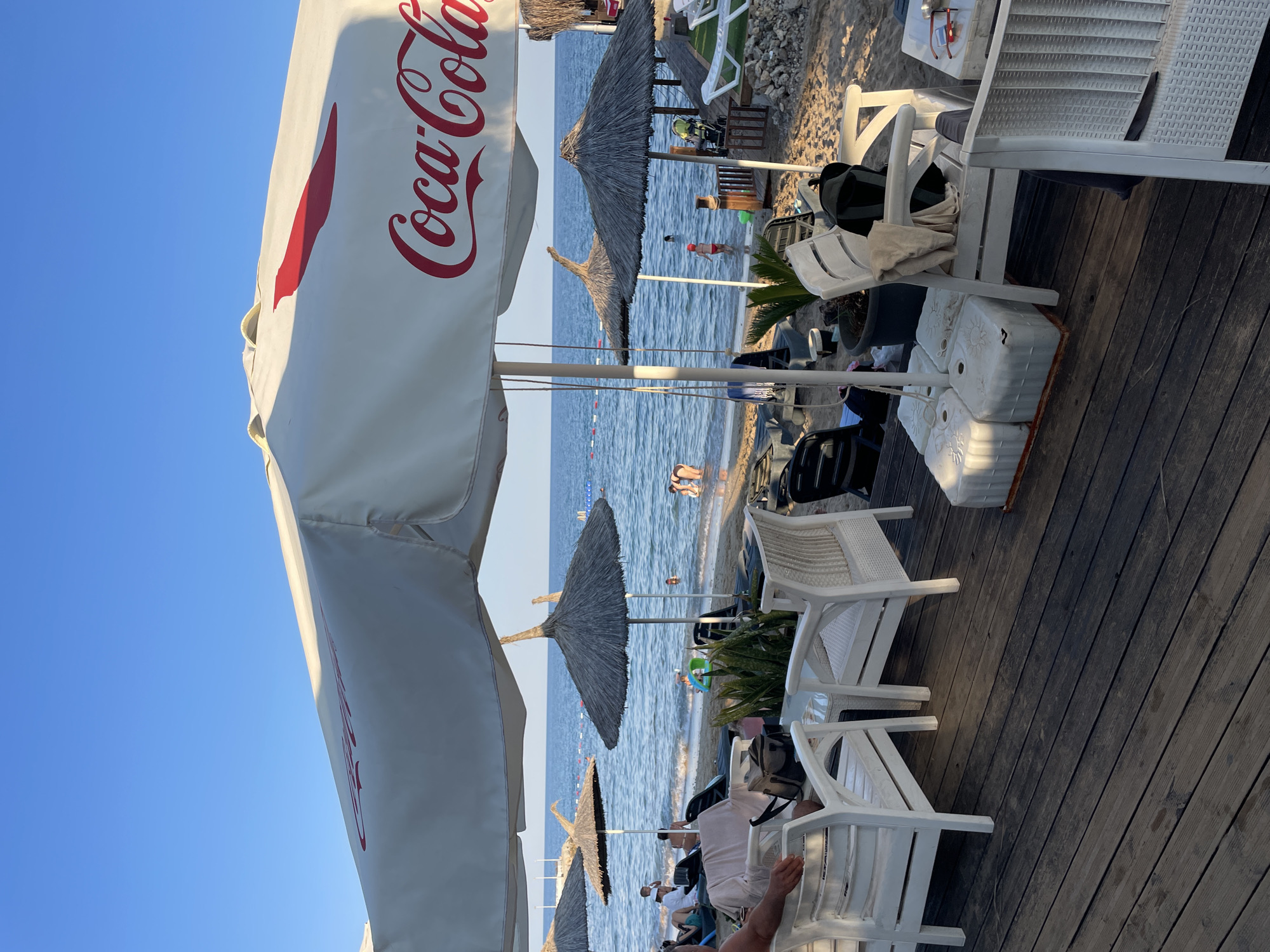 A Coca-Cola branded umbrella shades a wooden deck area with white seating and tables. Beyond the deck, a sandy beach is visible with thatched umbrellas, lounge chairs, and people in the shallow water of the Mediterranean Sea. The sky is clear and blue. The scene appears to be a beachside cafe or resort in Trikomo, Northern Cyprus.
