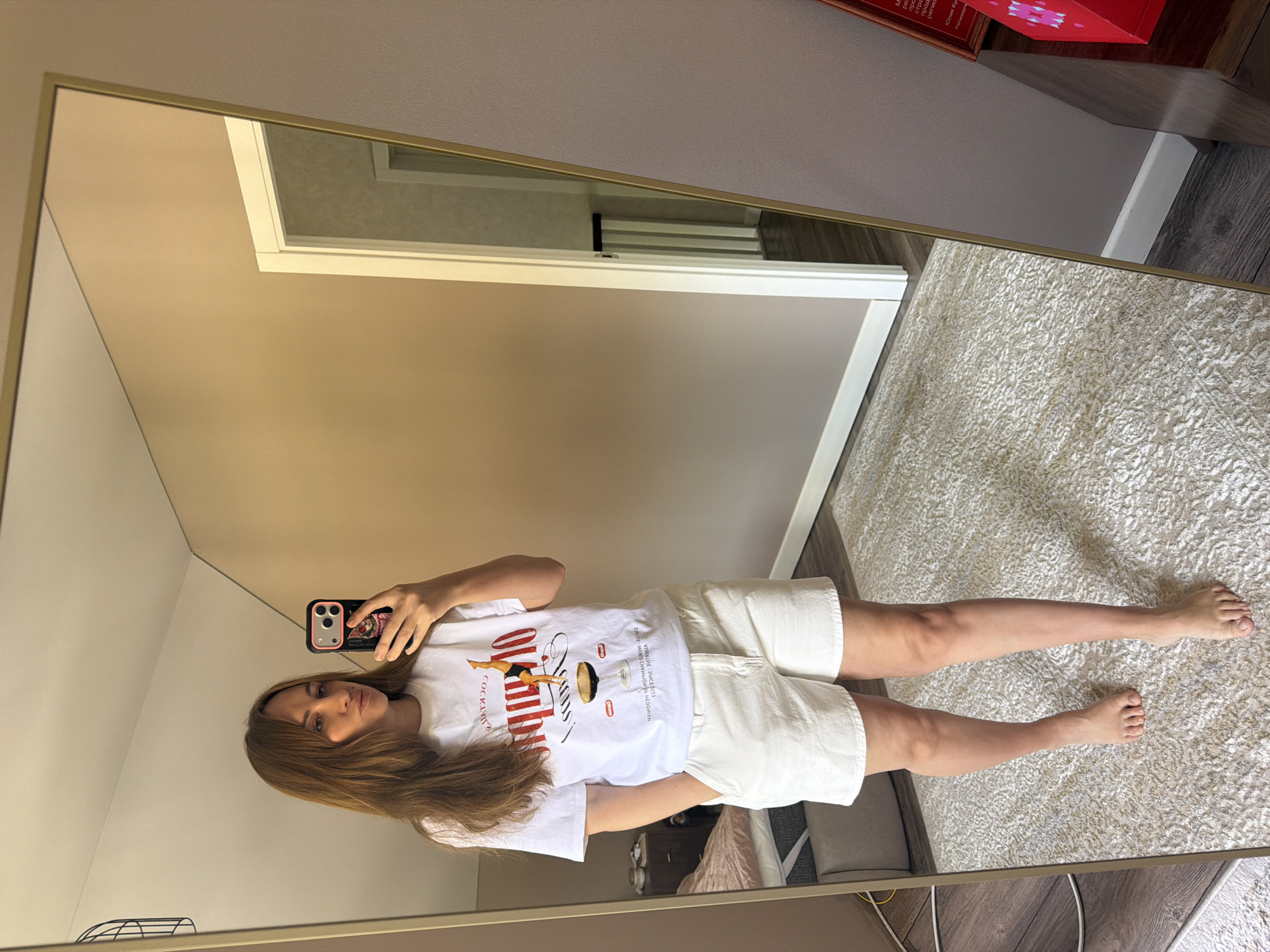 A young Caucasian woman with shoulder-length brown hair stands in a bedroom, taking a mirror selfie. She is wearing a white t-shirt with a graphic print of a cocktail and the word "COCKTAILS" in red, and white denim shorts. Her left hand is in her pocket. The room has light brown walls, a white rug, and a wooden floor. In the reflection of the mirror, a doorway leads to a hallway with a staircase. The woman is barefoot. The scene takes place in the city of Malinovka, Russia.