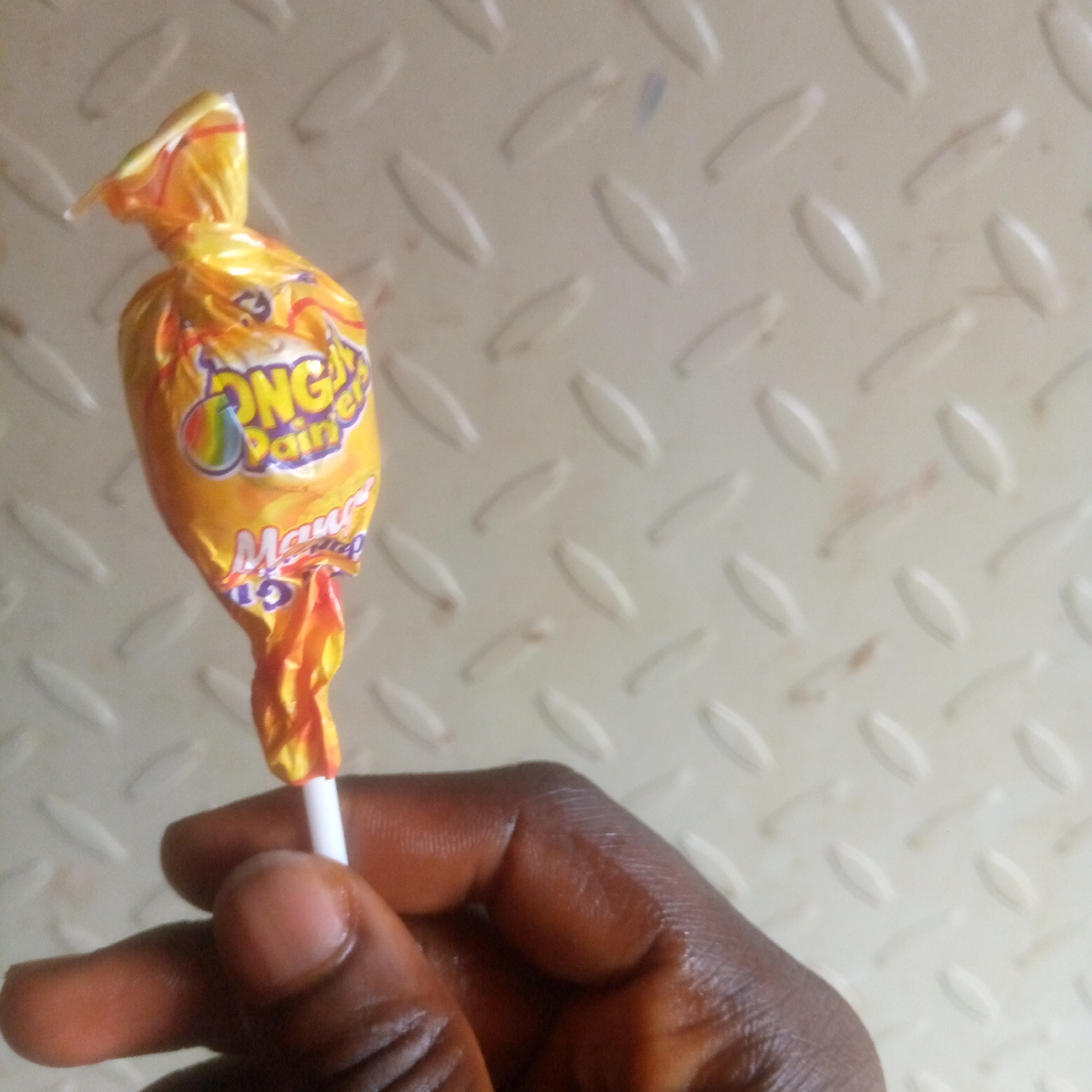A close-up, medium shot captures a hand holding a wrapped lollipop. The lollipop has a yellow and orange wrapper with colorful lettering that appears to say "ONG Y PAIN ER" and "Mango." The background is a light-colored, textured surface with diamond-shaped patterns. The hand, with dark skin tone, is holding the white stick of the lollipop between its thumb and index finger. The focus is on the lollipop and the hand, with the background slightly blurred. The scene appears to be indoors or in a shaded outdoor area, with neutral lighting. The location is Unknown, Unknown.
