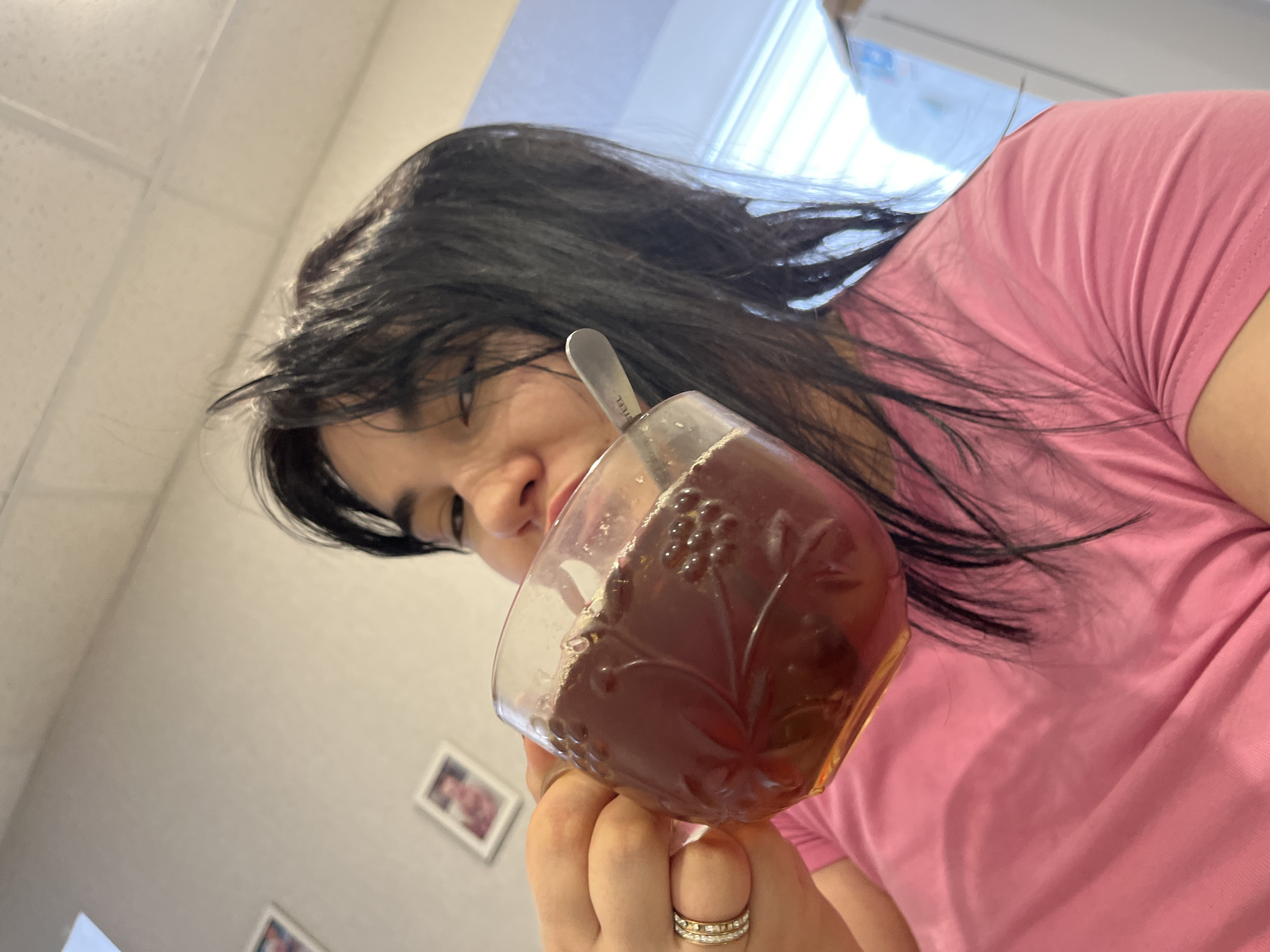 A woman is holding a glass of dark-colored liquid with a spoon inside. She is wearing a pink t-shirt and multiple rings on her fingers. The background includes a white ceiling, a window with blinds, and framed pictures on a white wall. The location is indoors, possibly in a residence. The city of Chaikovsky, Russia cannot be confirmed based on the visual information provided.