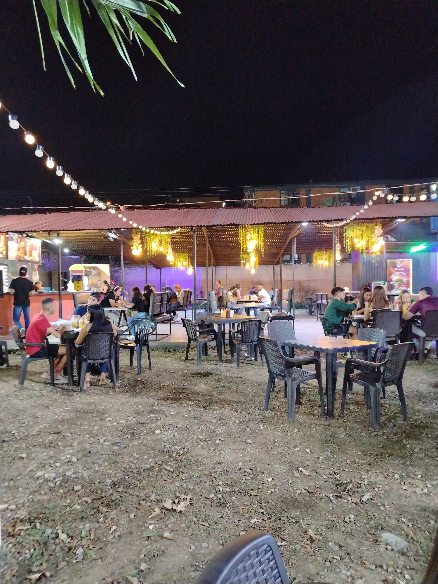 The image depicts an outdoor dining area in Tinaquillo, Venezuela at night. Several groups of people are seated at black plastic tables and chairs, engaged in conversation and eating. String lights hang above the tables, providing illumination along with other light sources. Underneath the lights and above the tables, decorative hanging plants are visible. The ground is dirt and gravel, scattered with leaves. A covered structure provides shelter for the dining area. In the background, a building can be seen. A food stand is also present to the left with a person standing behind it.
