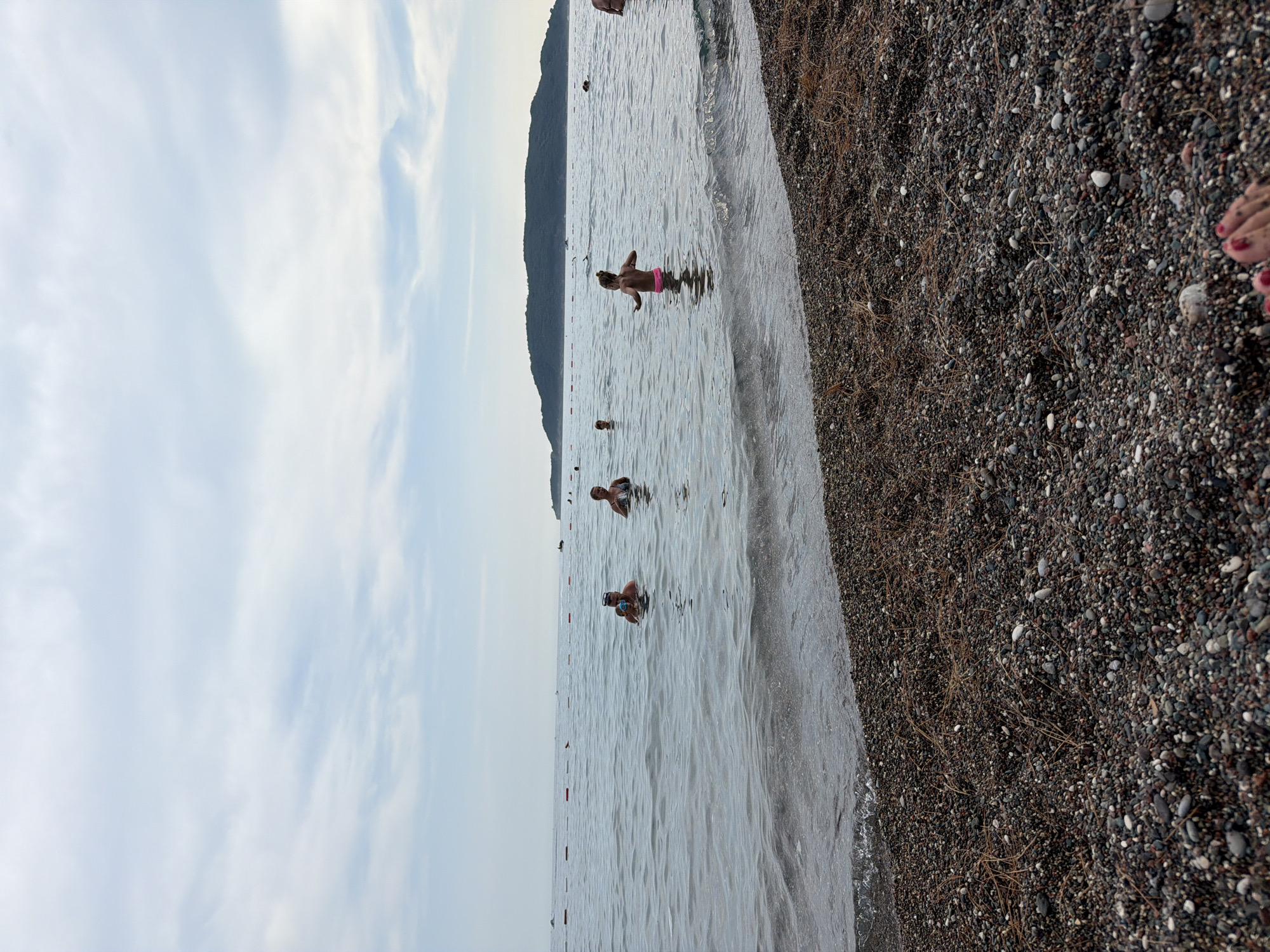 The image depicts a beach scene in Rafailovići, Montenegro. The camera is oriented vertically, showing a sliver of the beach in the foreground and the sea stretching towards the horizon. The beach is covered in dark pebbles and some dry vegetation. In the water, several people are visible, appearing to be enjoying a swim. One person, wearing pink swimwear, is standing in the shallow water, facing away from the camera with arms outstretched. Further out, other individuals are swimming or wading. The sea is calm with gentle waves lapping the shore. In the distance, a mountainous silhouette is visible against a pale, cloudy sky. The lighting suggests it is daytime, possibly late afternoon, given the soft light. The overall mood is serene and recreational, with people engaged in typical beach activities. There is no visible text in the image.