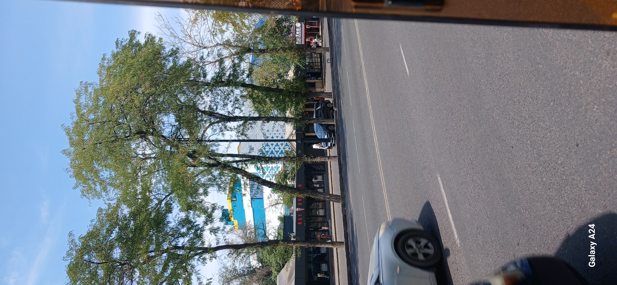 In Bishkek City, Kyrgyzstan, a street view captures a sunny day with a clear blue sky. Tall trees with green foliage line a sidewalk in front of buildings. One building features a distinctive modern architectural design with blue and yellow sections. On the street, a silver car is in motion on the left, and a dark SUV is parked on the right. People are visible on the sidewalk, some near storefronts. The overall scene depicts an urban environment with commercial establishments and residential structures, typical of a city in Central Asia.