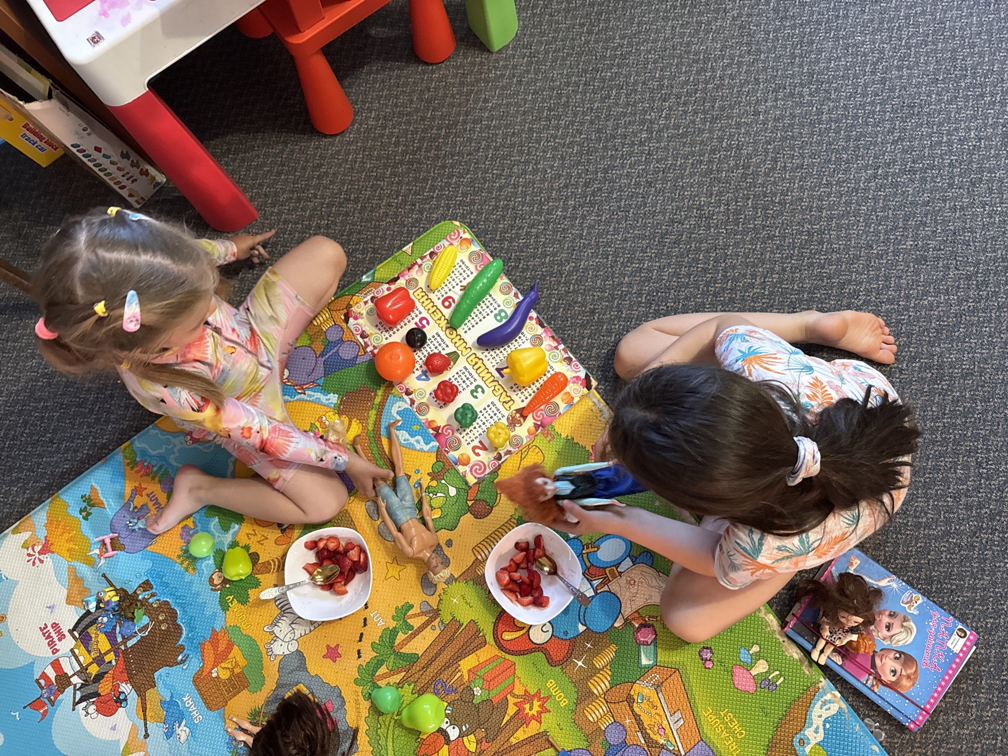 This is a high-angle, full shot of two young girls playing on a colorful play mat on a carpeted floor. The play mat features cartoonish illustrations of animals, a pirate ship, and various objects. The girls are dressed in casual, summer-themed clothing. The girl on the left has blonde hair with colorful barrettes and is wearing a long-sleeved swimsuit. She is reaching towards the play mat. The girl on the right has dark hair tied back with a scrunchie and is wearing a short-sleeved top. She is holding a toy and appears to be interacting with a doll.
Scattered on the play mat are several bowls of sliced strawberries, spoons, a few plastic toy fruits and vegetables (an eggplant, a bell pepper, corn, and an apple), and two dolls. One doll is a male figure lying on the mat, while the other is a female doll held by the girl on the right. A book with illustrations of characters from the movie "Frozen" is also on the mat near the girl on the right. In the background, partially visible, is a red and white child's table or play structure.
The overall scene suggests a playdate or a quiet playtime session. The lighting appears to be indoor, with no clear indication of the time of day or weather. The text visible includes "TREASURE CHEST," "BOMB," "SHARK," and "PIRATE SHIP" on the play mat. Additionally, there is text on a small booklet or card that appears to be a multiplication table in Ukrainian, with phrases like "ТАБЛИЦЯ МНОЖЕННЯ" and some math equations. There is also text on a box in the background that reads "Building block track car."