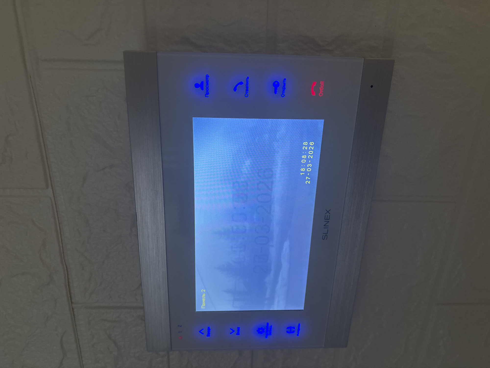 A SLINEX video intercom system is mounted on a light-colored, textured wall, resembling decorative faux brick or stone paneling, in the city of Ферма, Russia. The device features a central blue-tinted screen displaying a pixelated, out-of-focus image of what appears to be a natural landscape with trees or a forest. Overlaid text on the screen includes "Панель 2" (Panel 2) in the top left corner, and "18:08:28 27-03-2026" in the bottom right, indicating the time and date. Fainter, larger text "20-03-202X" is partially visible in the screen's center. To the left of the screen are four vertically arranged, blue-illuminated capacitive buttons with generic icons (up arrow, down arrow, gear, house). To the right are three vertically stacked, blue-illuminated capacitive buttons labeled in Cyrillic: "Просмотр" (View) with a person icon, "Ответить" (Answer) with a phone receiver icon, and "Открыть" (Open) with a key icon. Below these is a single red-illuminated capacitive button labeled "Отбой" (Hang up) with a phone receiver icon. The wall texture is uniform across the visible area above and around the device.