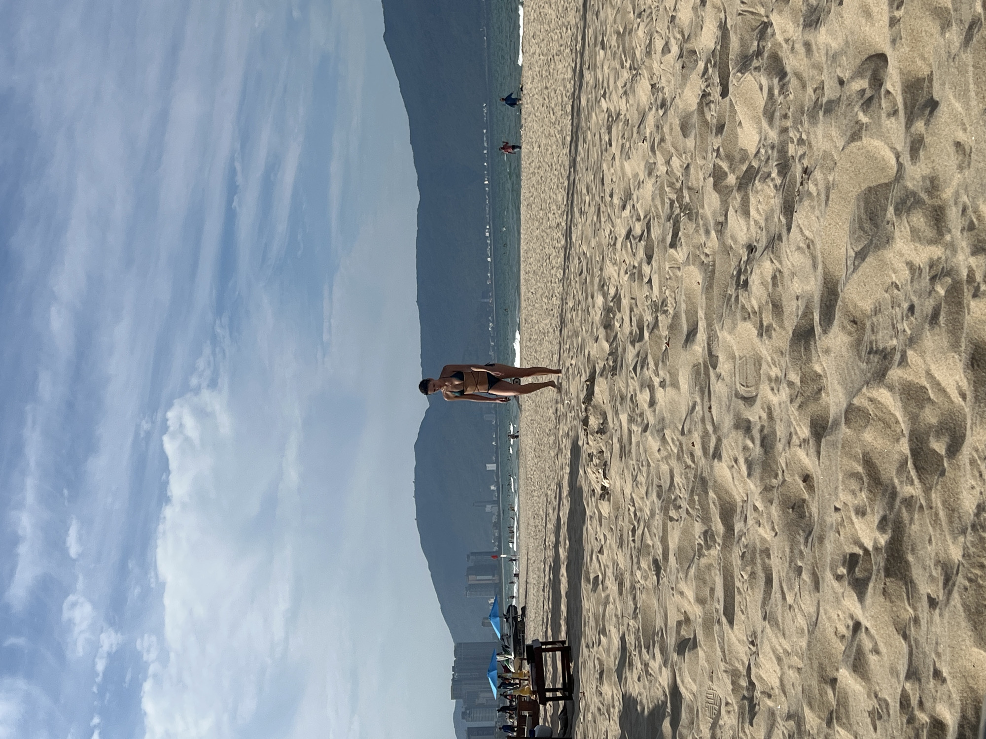 A woman in a dark bikini walks on a sandy beach towards the viewer, with the ocean and a mountainous city skyline in the background. The sky is partly cloudy. In the mid-ground, there are beach umbrellas, lounge chairs, and people in the water and on the shore. The foreground is dominated by textured sand dunes. The scene appears to be on My Khe Beach in Đà Nẵng, Vietnam, given the presence of the distinct Marble Mountains in the distance and the general appearance of the coastline.