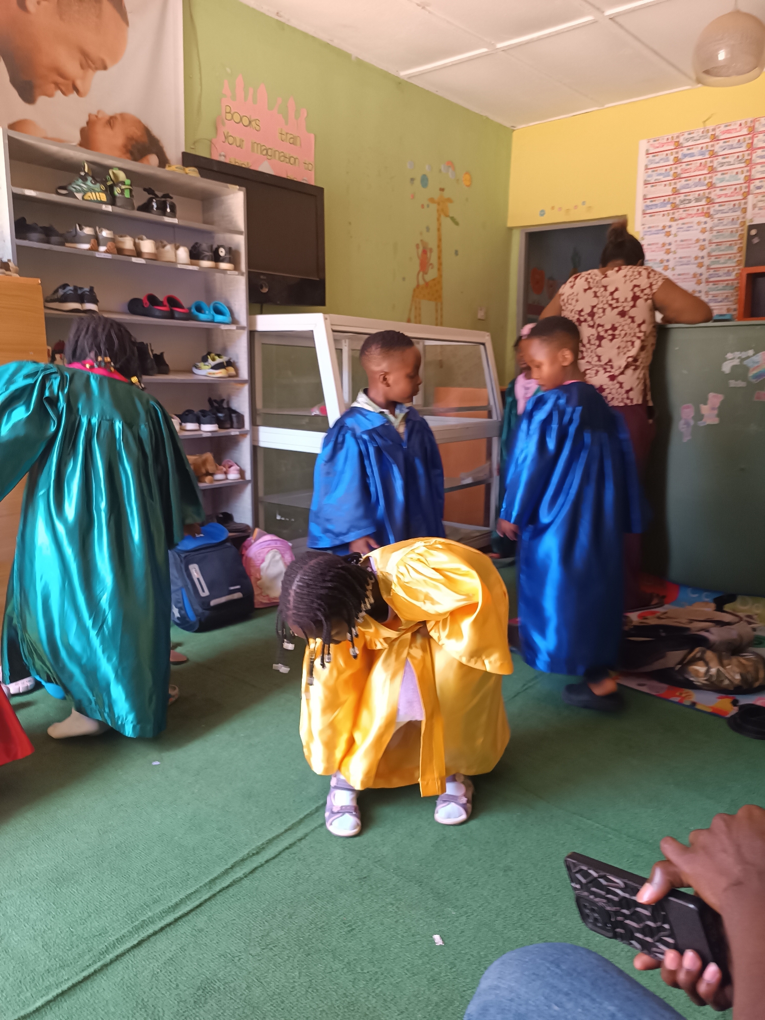 The image depicts an indoor scene that appears to be a classroom or a children's play area, likely located in Jos, Nigeria. In the foreground, a young child wearing a yellow graduation gown is bent over, with their back to the camera. To their left, another child in a bright green graduation gown stands partially visible. To the right and further back, two other children are dressed in blue graduation gowns. A woman, presumably a teacher, is standing with her back to the camera, dressed in a patterned top and dark skirt, near a counter or desk. The room has brightly colored walls with children's artwork, including a giraffe poster. A shelf unit filled with shoes is visible on the left. Backpacks are placed on the floor near the children. The flooring is a green carpet with a distinct cord or seam running across it. In the lower right corner, a person's hand is holding a black mobile phone. The overall impression is of a celebratory event, such as a graduation ceremony, for young children.