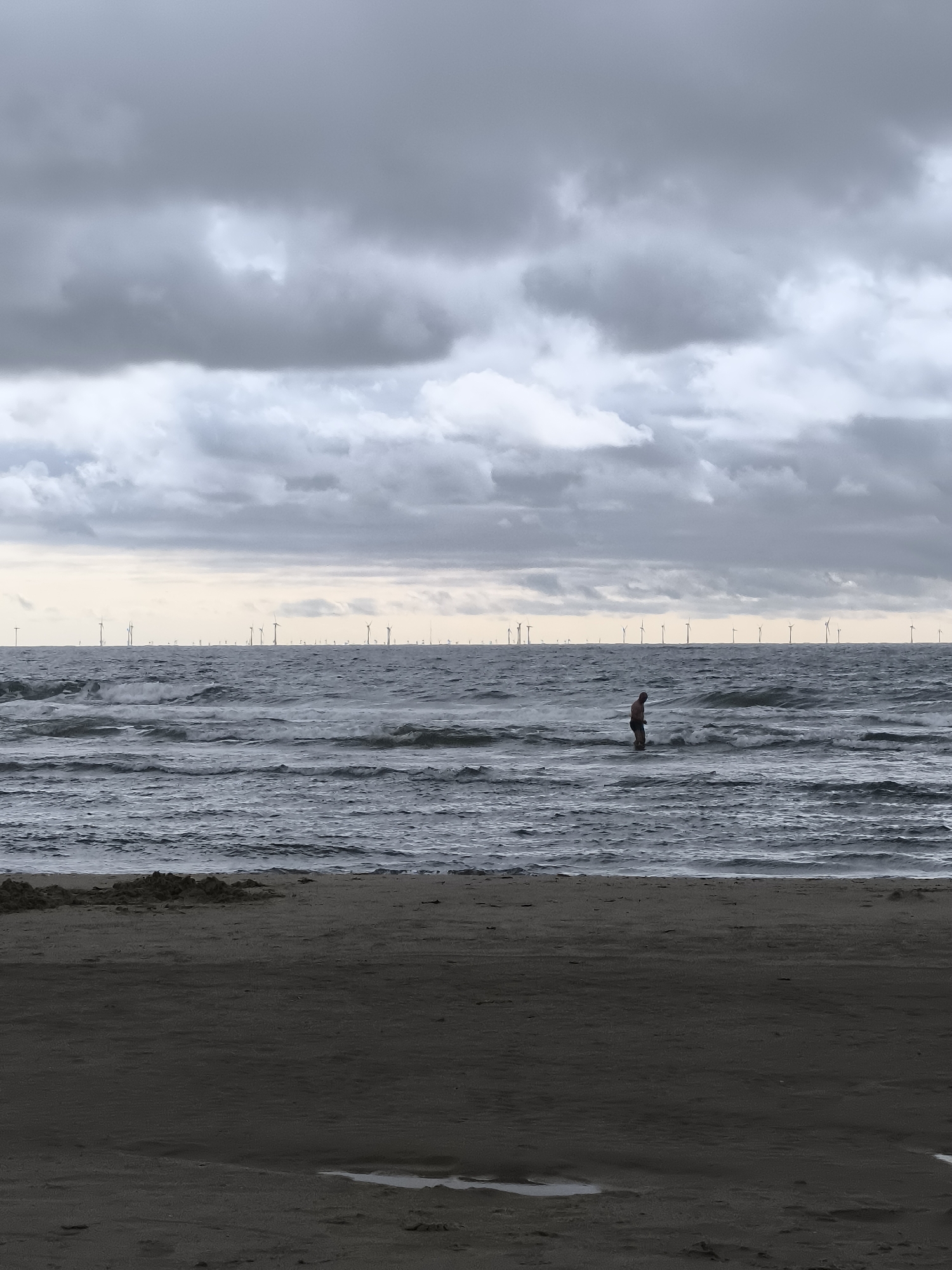 A solitary person stands in the choppy, grey sea, facing away from the camera. The water is dark and restless, with waves breaking around their knees. The beach in the foreground is dark and damp, with some small mounds of sand. In the distance, a vast wind farm stretches across the horizon, marked by numerous white turbine structures. The sky is overcast with heavy, dark grey clouds, with a pale sliver of lighter sky near the horizon. The scene evokes a sense of isolation and the power of nature. The image is likely captured on the coast near Bergen, Netherlands, given the presence of the offshore wind farm.